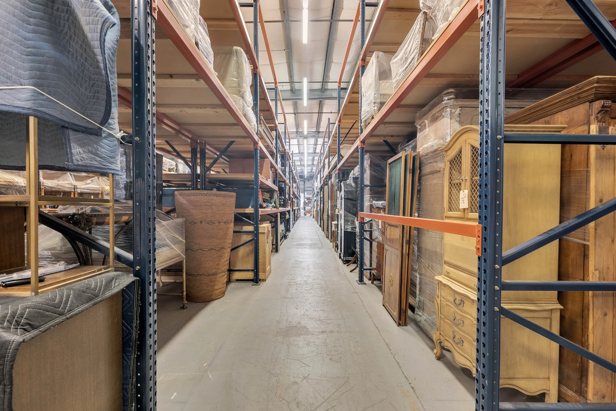 View of a warehouse aisle with metal shelving units holding furniture and other items, some wrapped in plastic.