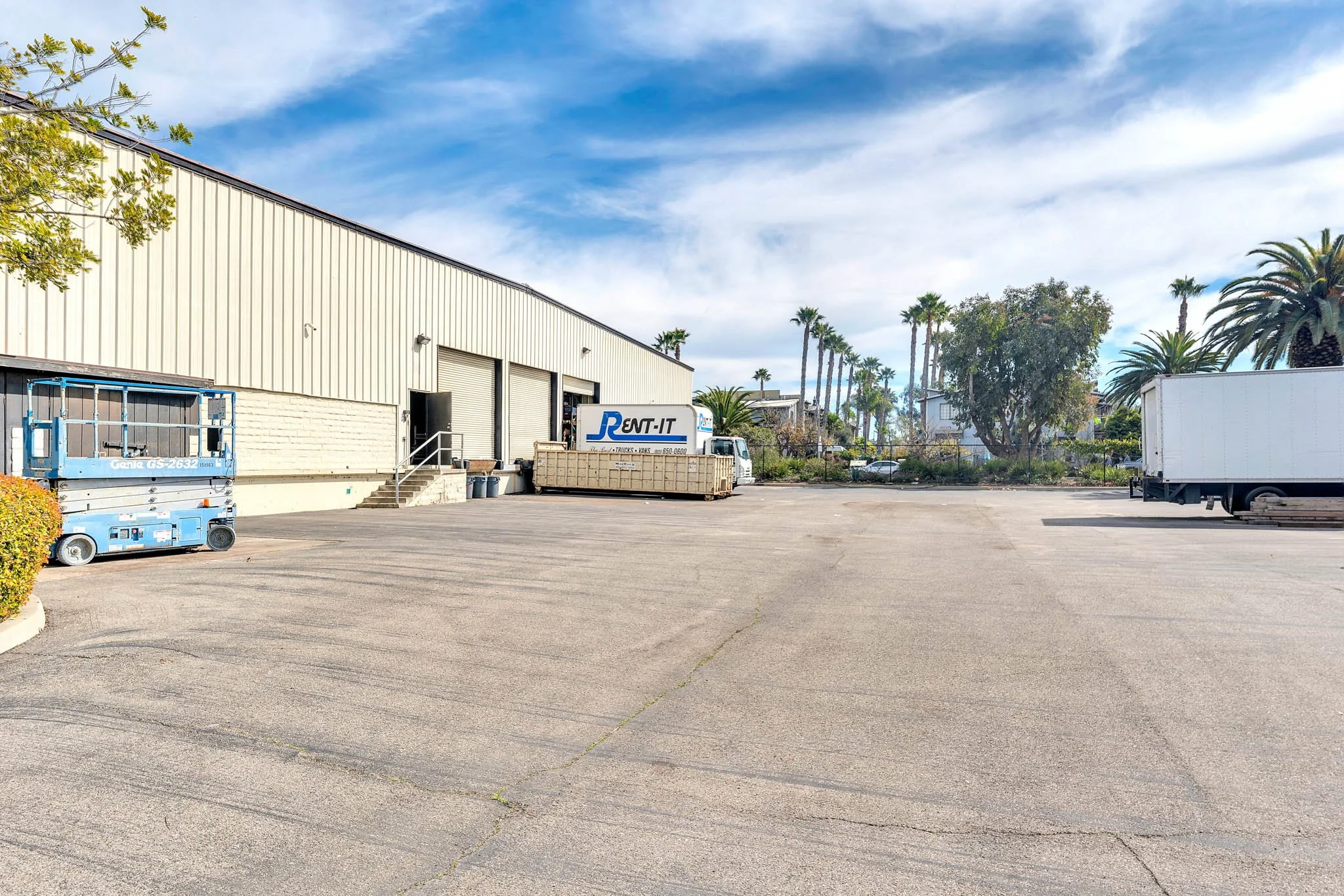 Empty parking lot outside a warehouse with trucks and equipment, surrounded by palm trees under a partly cloudy sky.