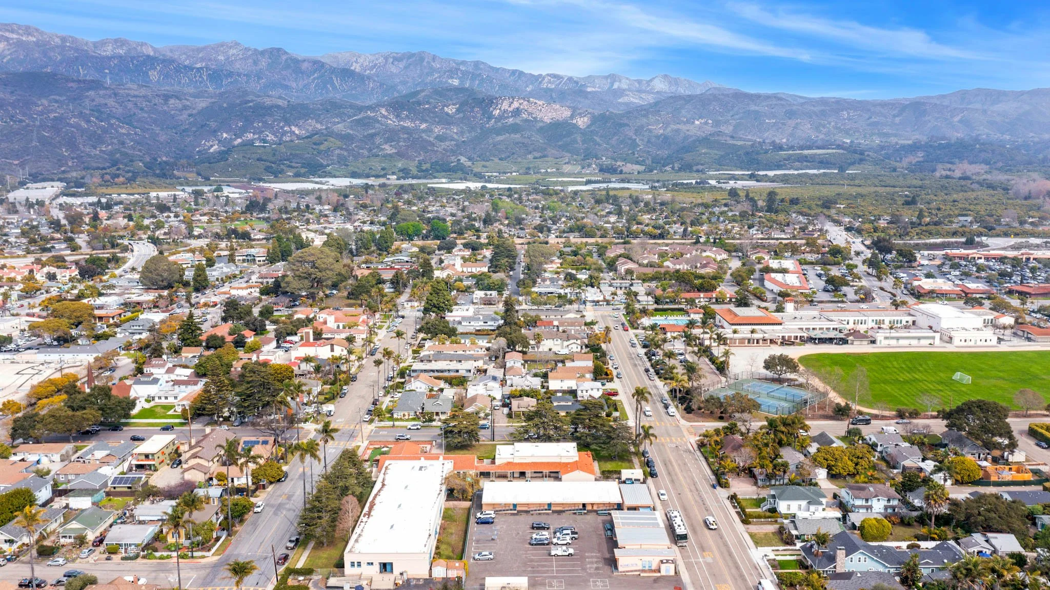 Aerial view of a suburban neighborhood with houses, trees, streets, a green sports field, and surrounding mountains under a blue sky