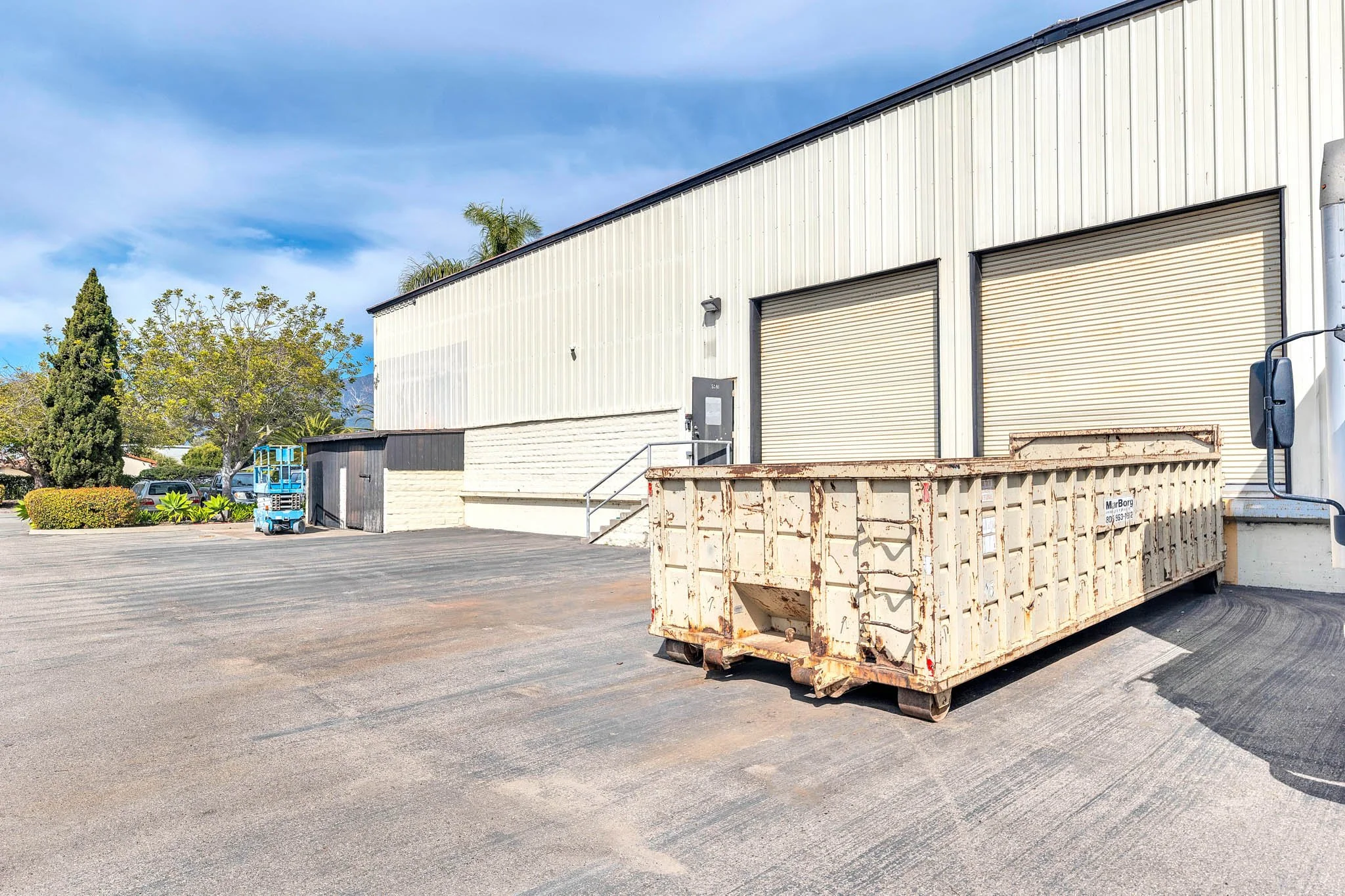 Industrial building with two closed roll-up doors, a rusty dumpster in the foreground, and parked cars and trees in the background on a clear day.
