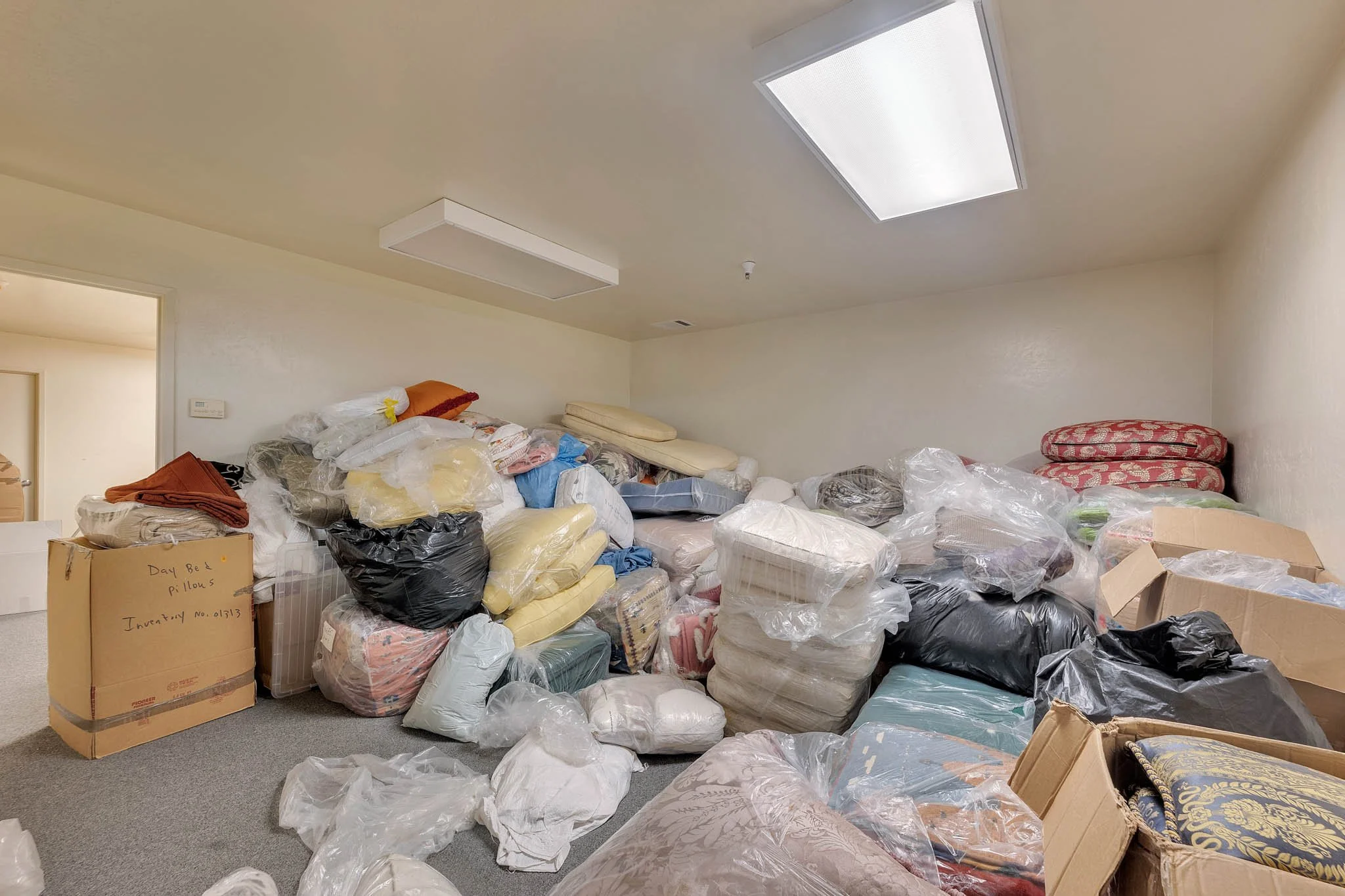 A cluttered room filled with piles of pillows, blankets, and plastic bags, with some pillows stacked on top of each other and some in open boxes.