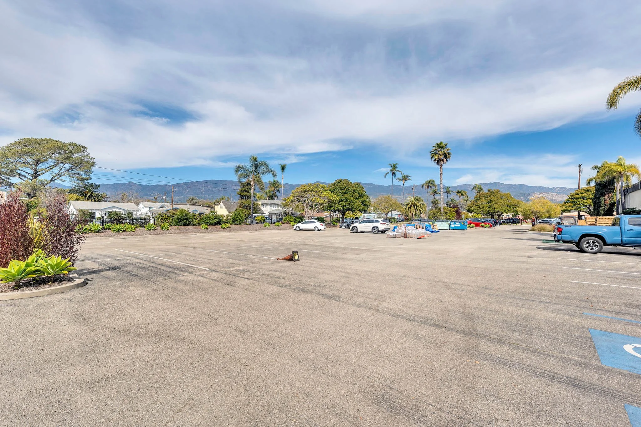 An empty parking lot under a partly cloudy sky with some vehicles, trees, plants, and mountains in the background.