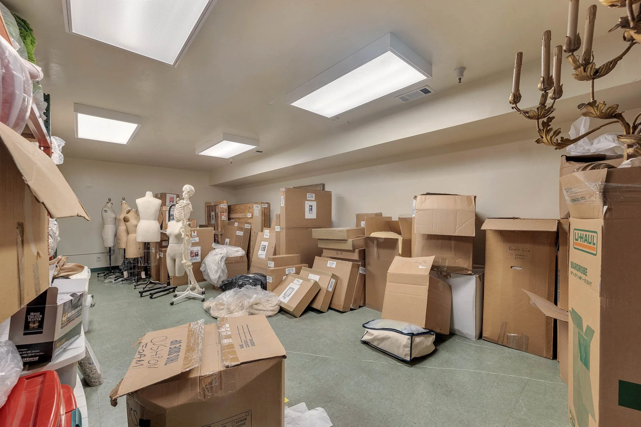 Storage room with cardboard boxes stacked against the wall, several mannequin torsos, and a small table on the left side. The room has a beige wall, green carpeted floor, and fluorescent ceiling lights.