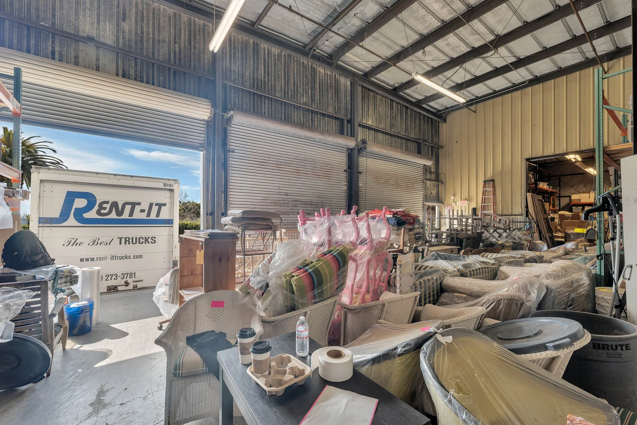 Interior of a warehouse or storage facility with furniture, boxes, and equipment, and a truck from Rent-It parked outside visible through the open door.