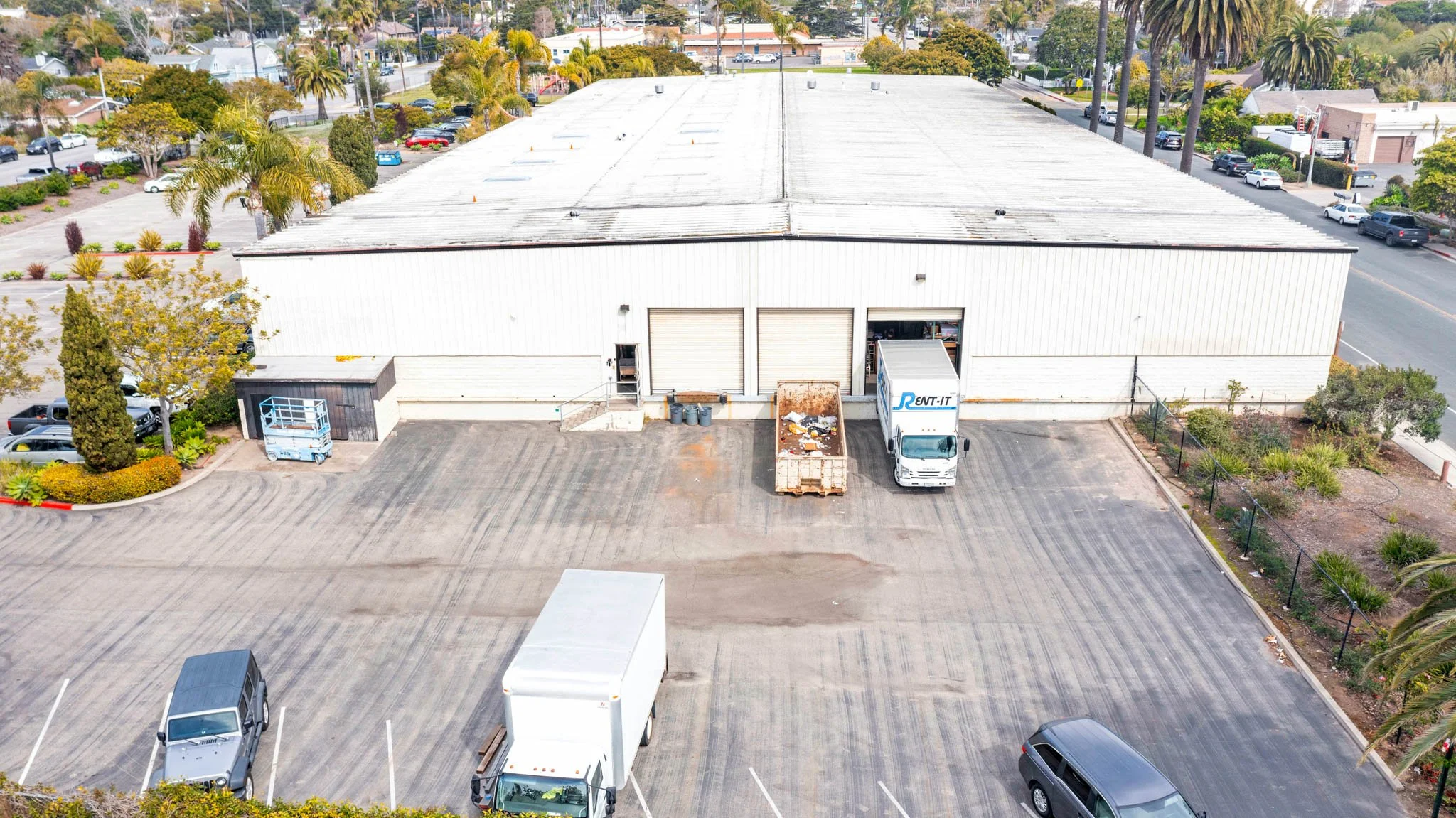 An aerial view of a large warehouse with a parking lot in front, several parked vehicles including trucks and cars, and some landscaping around the building's perimeter.