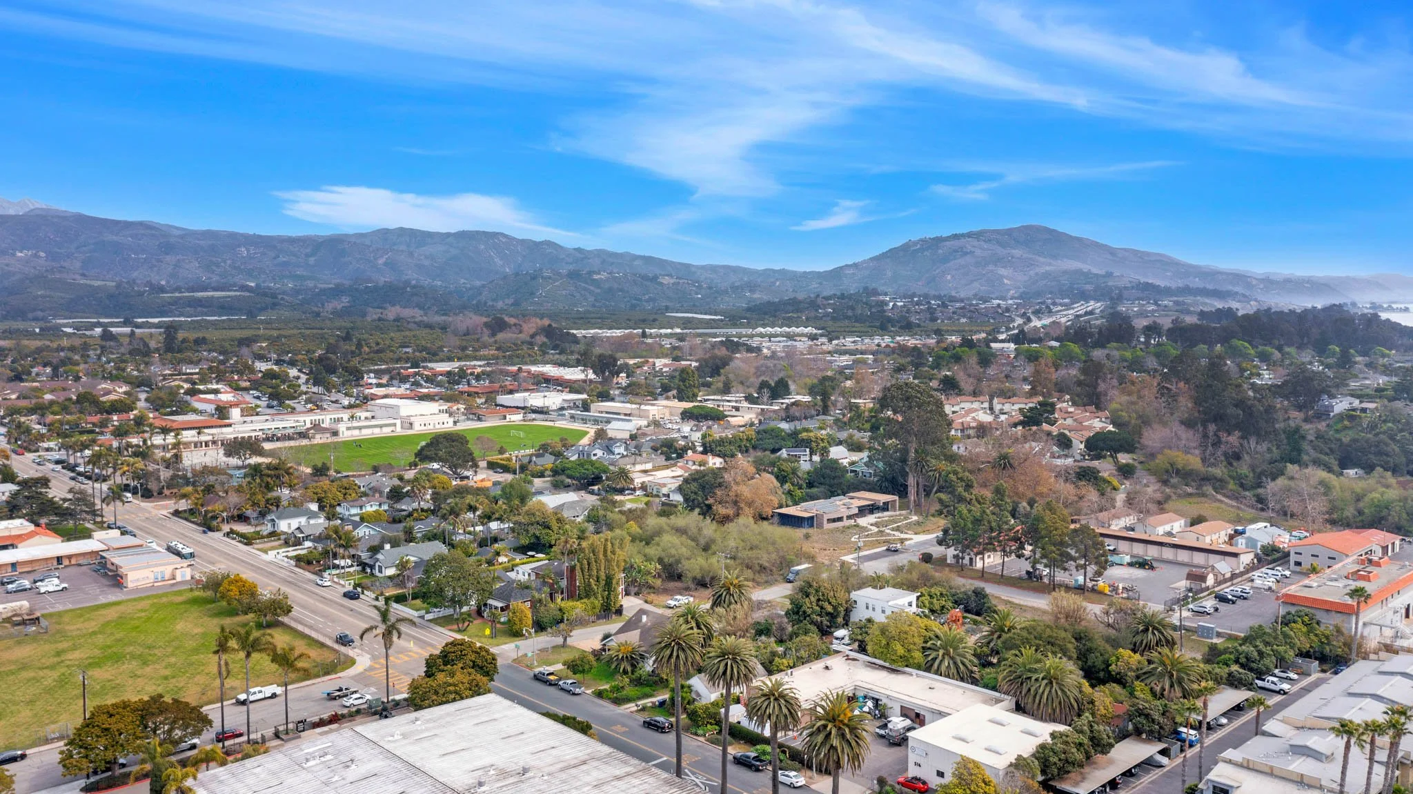 Aerial view of a suburban area with houses, trees, streets, a school with a green field, and mountains in the background under a blue sky.