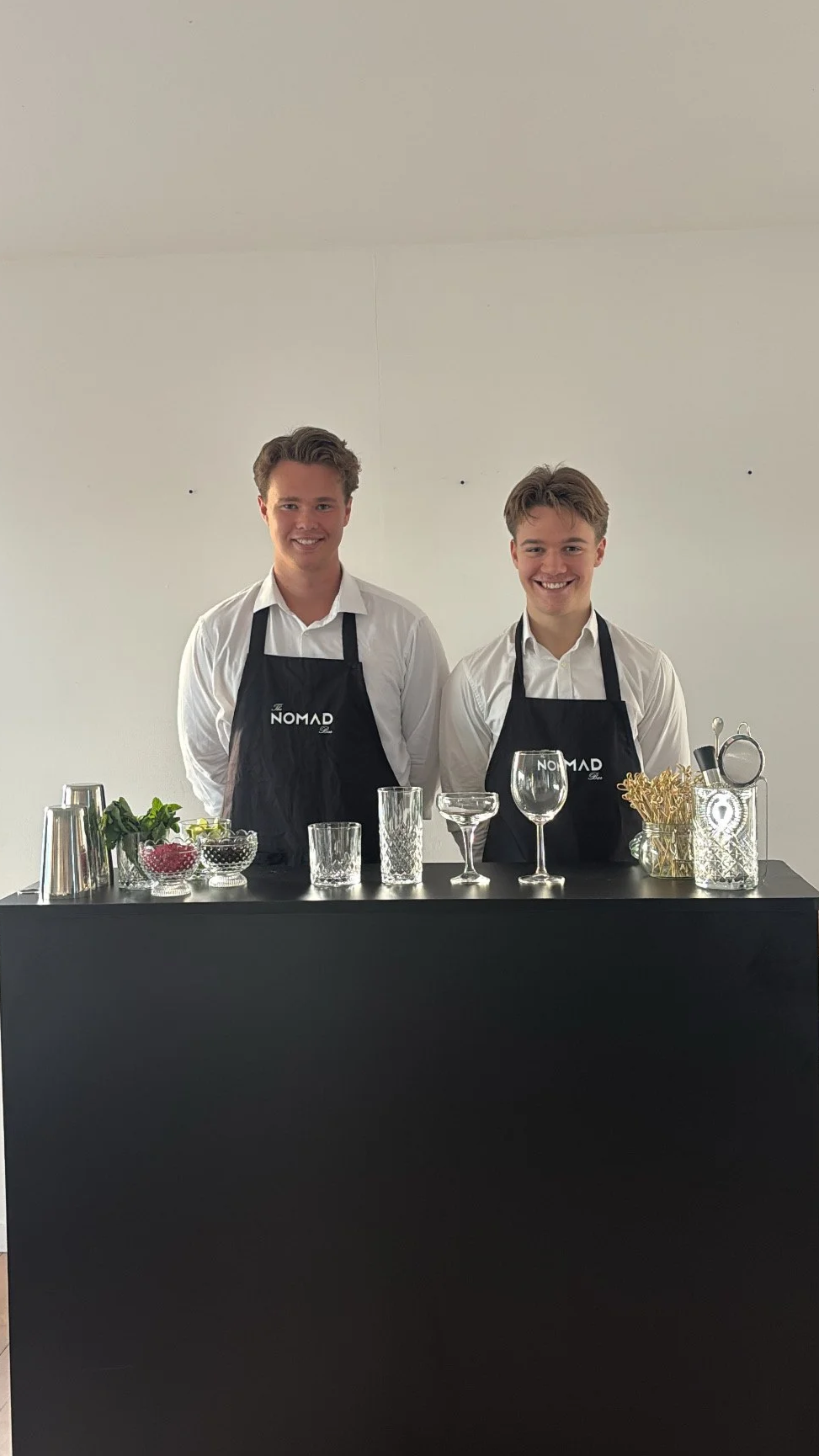 To unge mænd står bag en sort bar med forskellige cocktail-udstyr og ingredienser. Begge har hvide skjorter og sorte forklæder med 'NOMAD' logo.