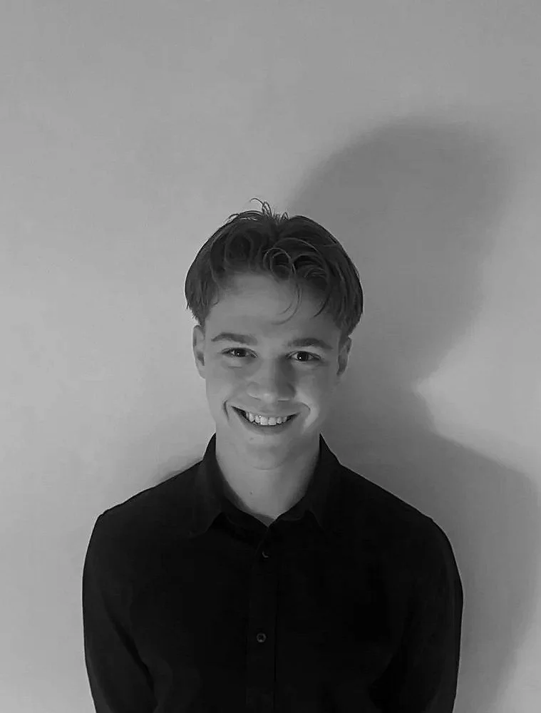Sort-hvid portrætfotografi af en smilende ung mand med kort, bølget hår iført sort skjorte, stående foran en neutral væg.