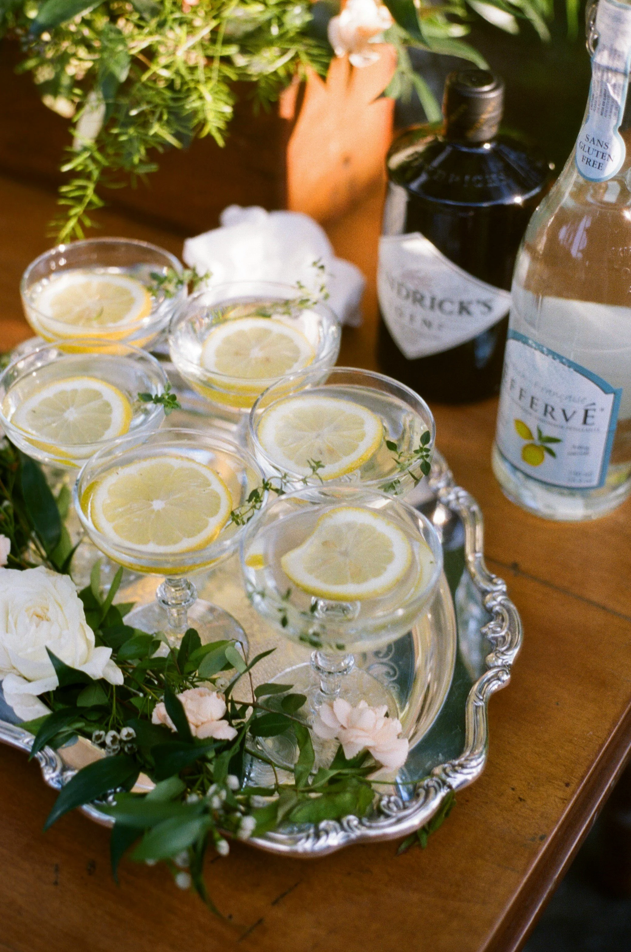 Bedukket bord med seks cocktailglas med citrusskiver, grønne urter, en flaske med Sprite, en flaske med tonic water, en kop med rom og nogle blomster og grønne planter i baggrunden.