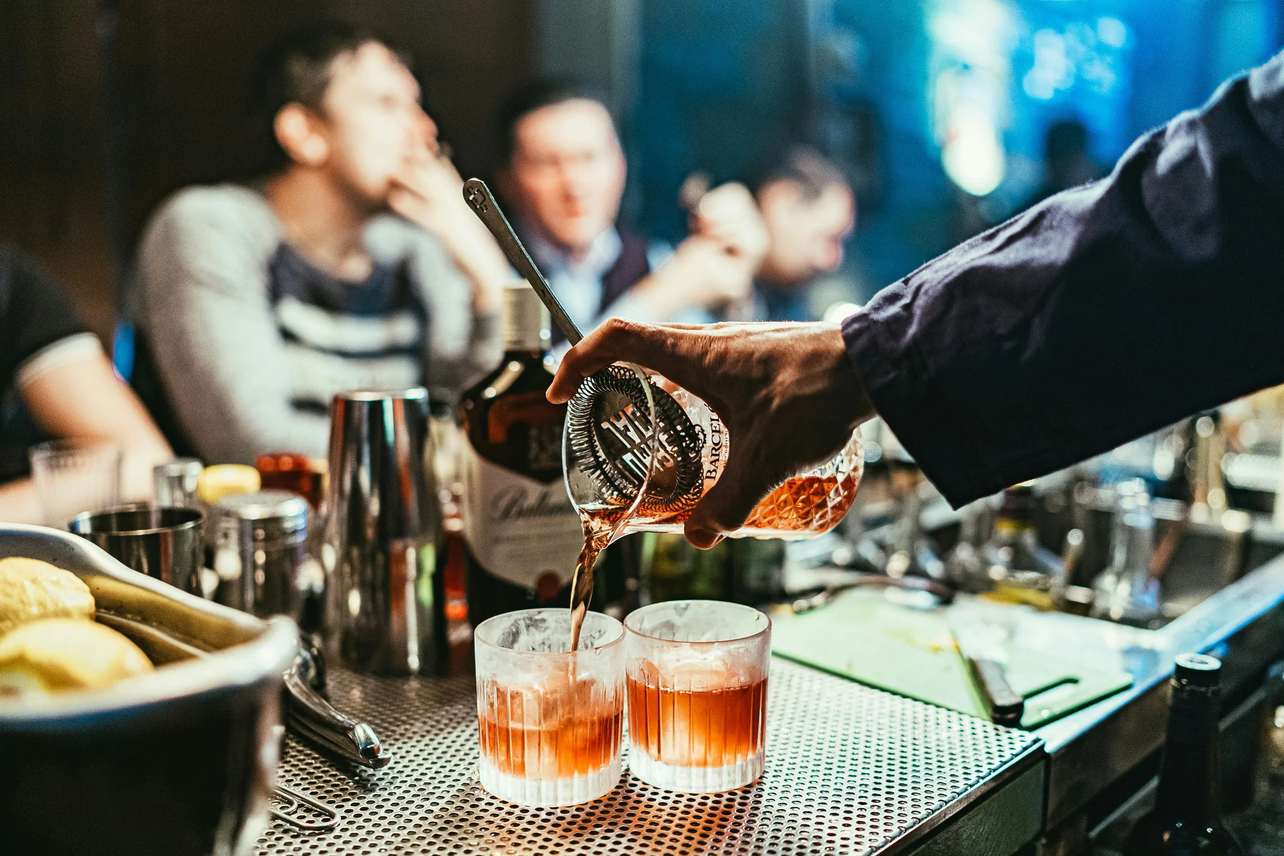 En person hælder en amberfarvet alkohol i to glas på en bar, mens andre mennesker sidder i baggrunden.