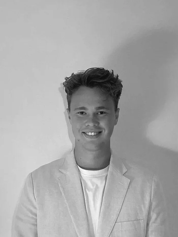 Et sort-hvide portrætbillede af en ung mand med kort, lyst hår, iført en lys blazer og en hvid t-shirt, smilende foran en blank væg.