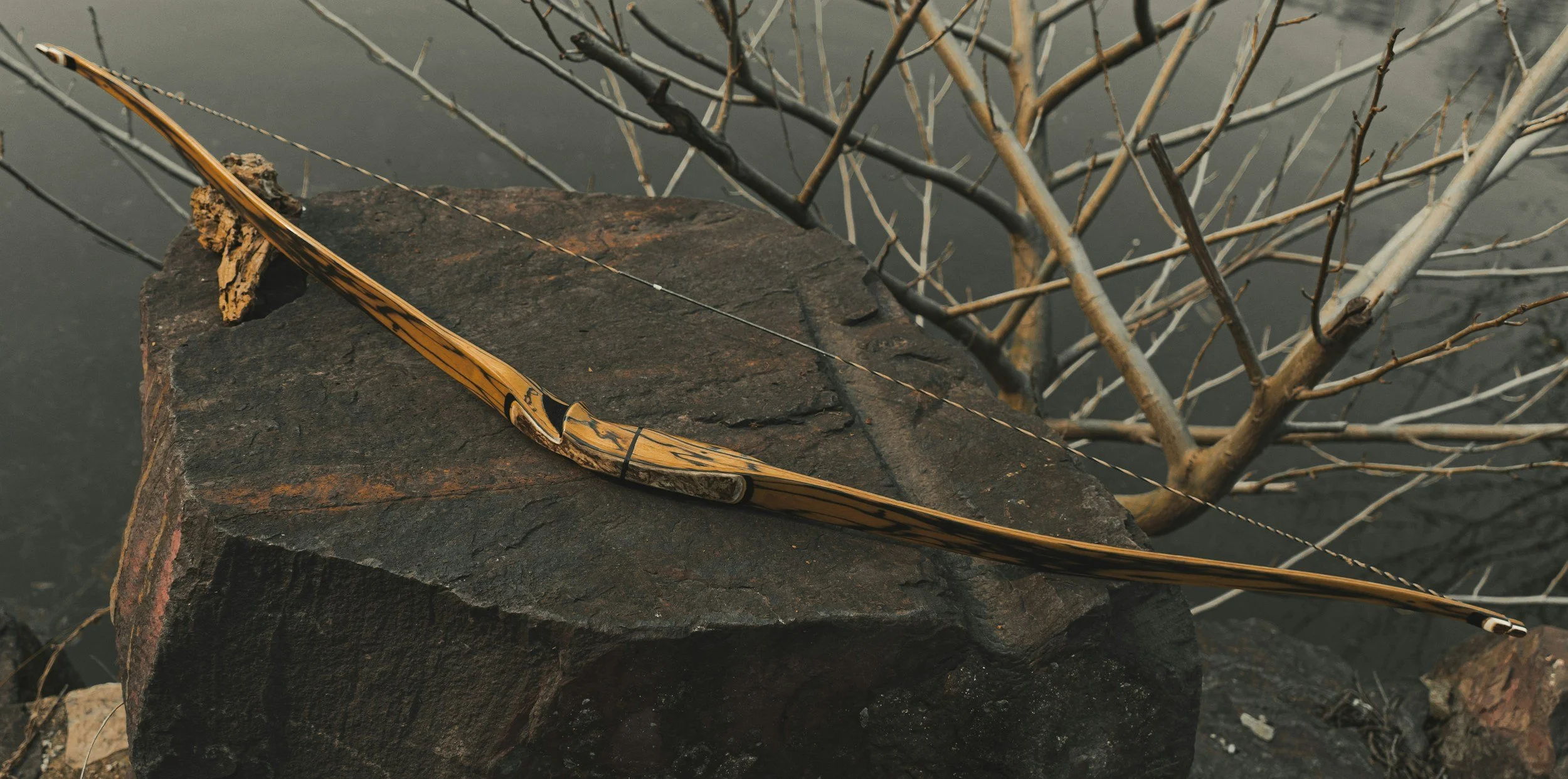 A wooden recurve bow resting on a large rock with a leaf attached, with bare tree branches in the background.