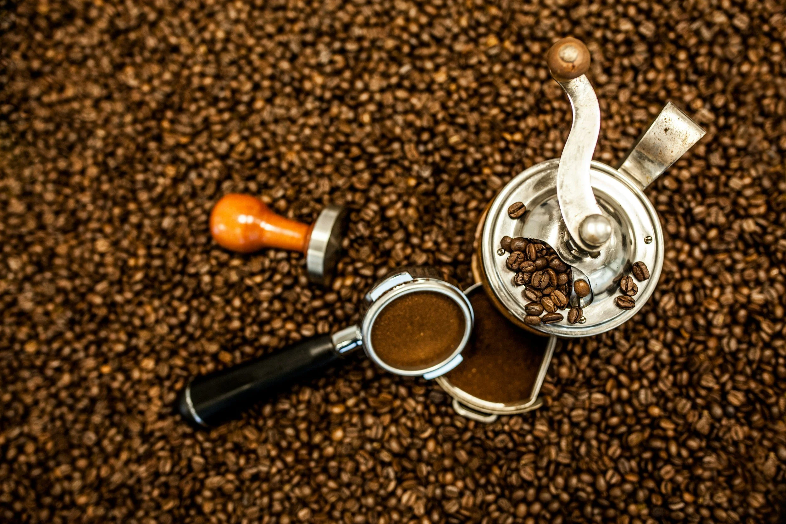 Coffee beans, a manual coffee grinder, an espresso shot, and a coffee tamper on a surface covered with roasted coffee beans.