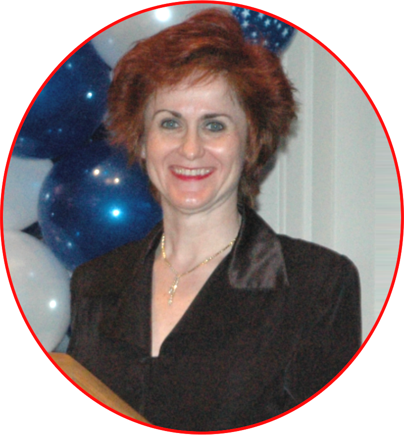 A woman with short, curly red hair smiling, wearing a black blouse with satin lapels and a gold necklace, standing in front of blue and white balloons.