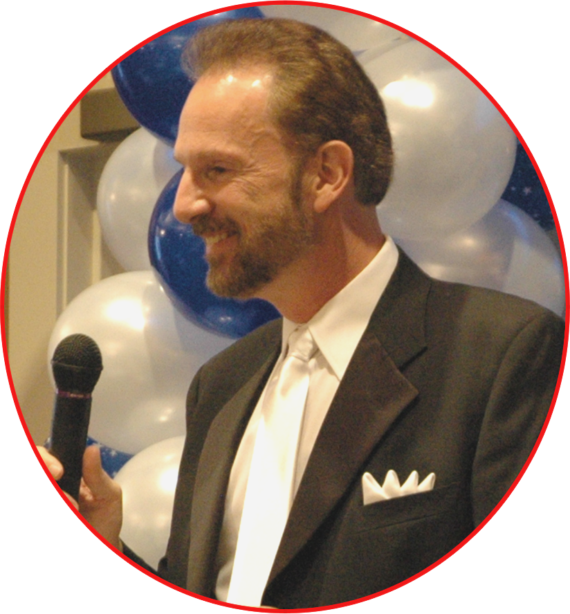 A smiling man in a tuxedo holding a microphone, standing in front of blue and white balloons.