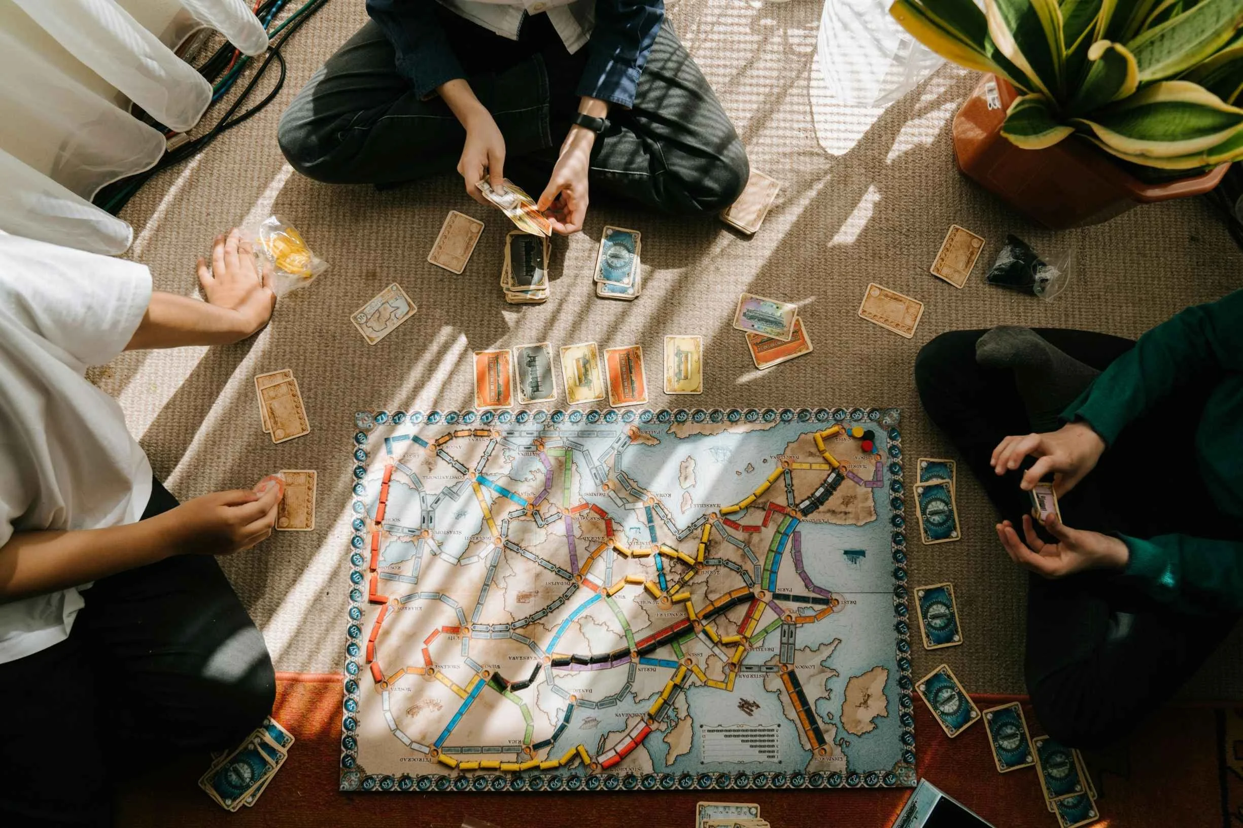 Three people are playing a board game, Risk, on a beige carpeted floor. The game board depicts a map of the world with various colored armies. Cards are scattered around the board and players are holding cards in their hands. One person's hand is rea