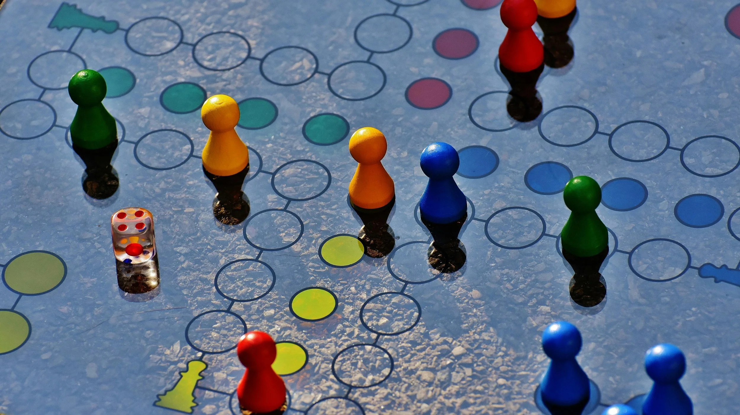 Board game with colored plastic pawns and a dice on a textured surface. The board has a network of circles and paths for game pieces.