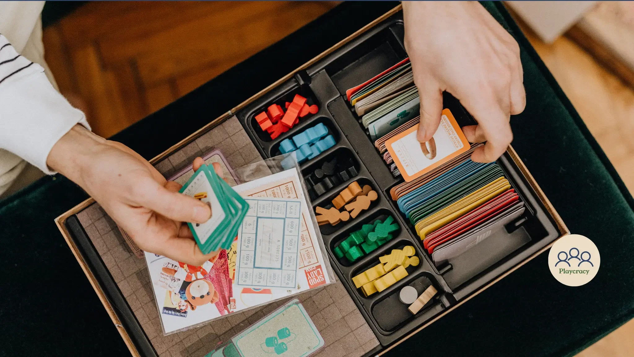 The components of a board game, with a hand holding some cards, and several multicolor meeples visible