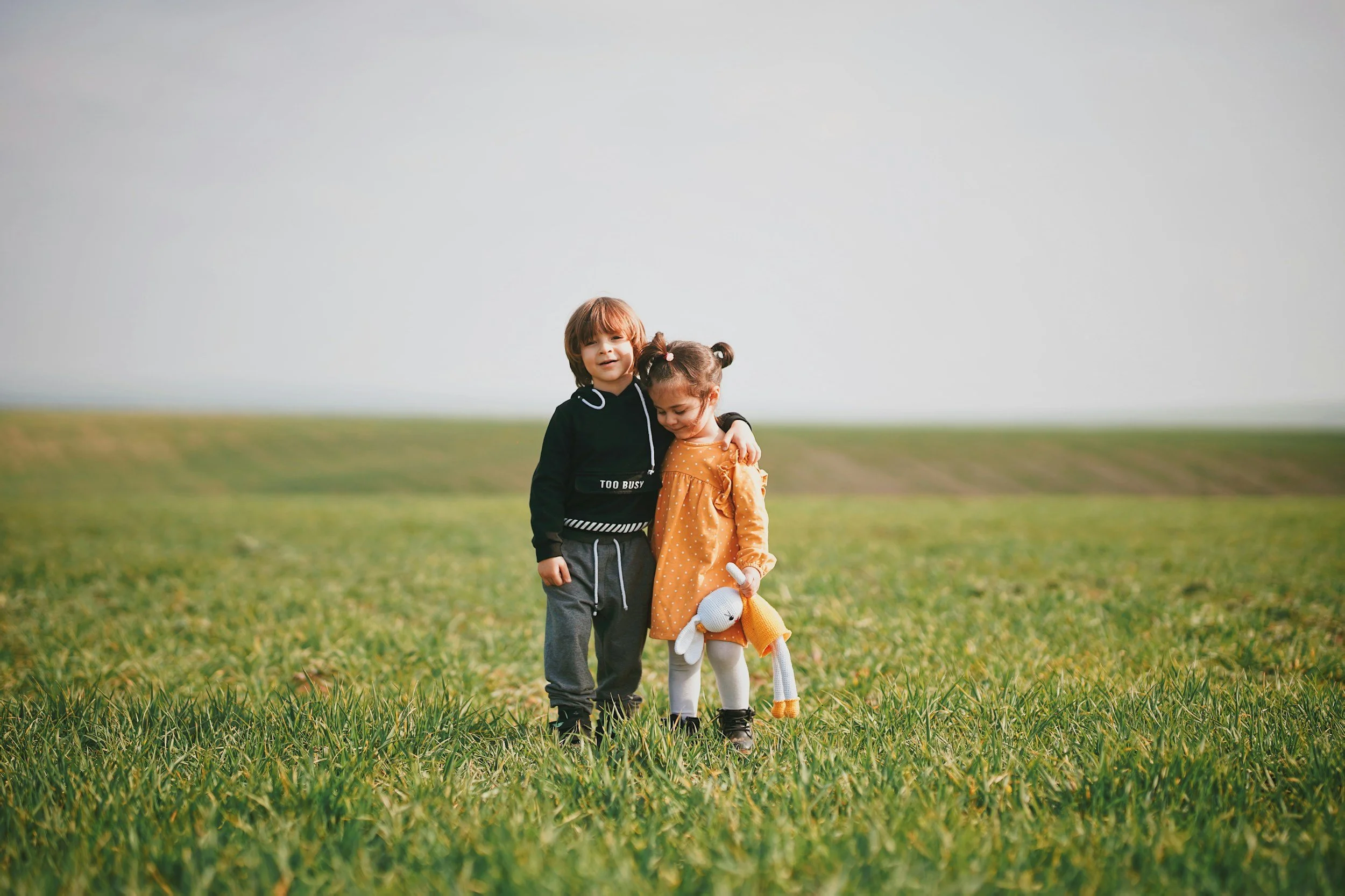 Two children, a boy and a girl, standing together in a grassy field with a blurry horizon and cloudy sky in the background. The boy is dressed in a black hoodie and gray pants, while the girl wears a yellow dress with a stuffed animal. They are hugging and standing close to each other.