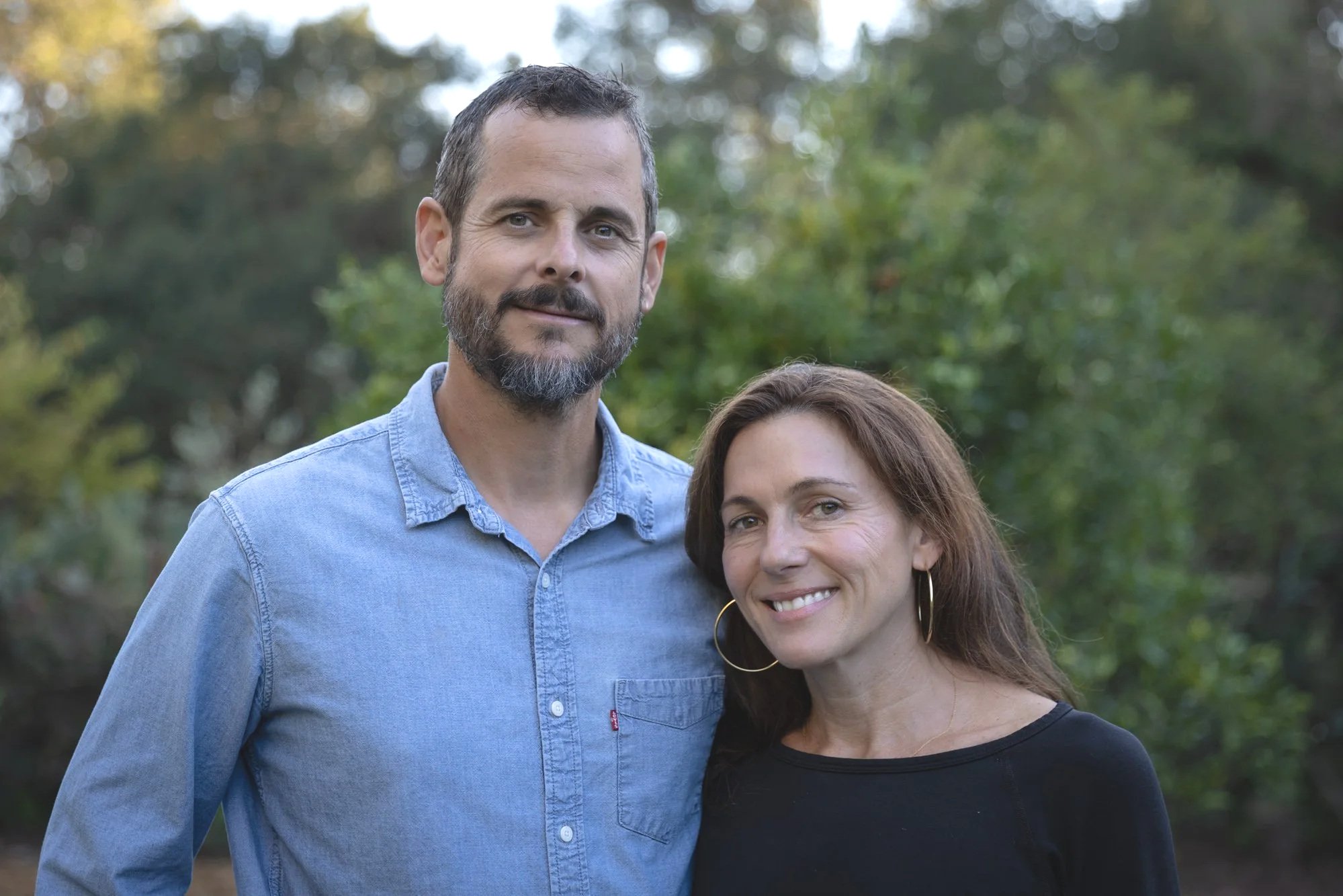 A man and woman standing outdoors in front of trees, smiling at the camera. The man has short dark hair and a beard, wearing a light blue denim shirt. The woman has long brown hair, wearing hoop earrings and a black top.