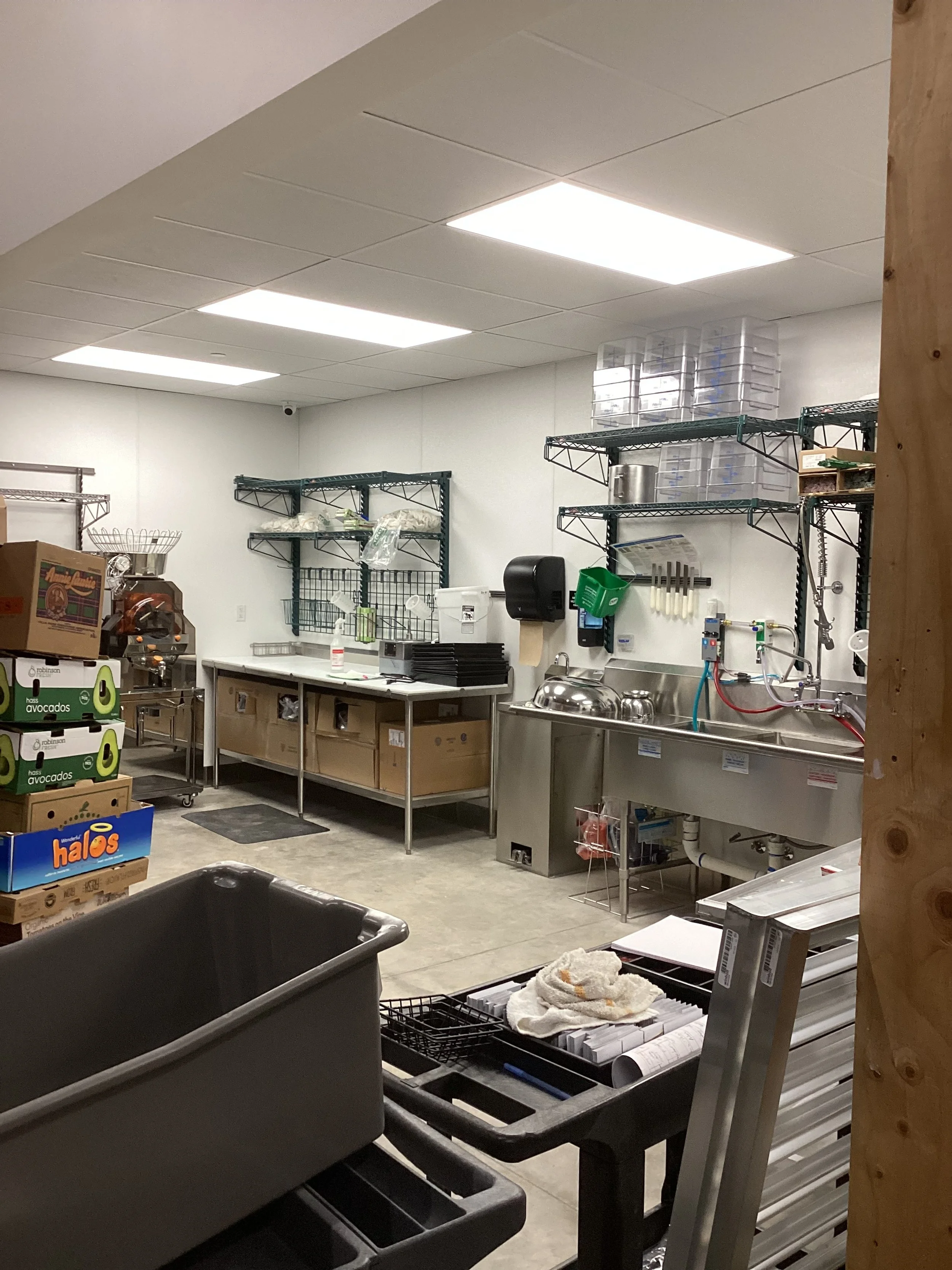 Commercial kitchen or food preparation area with stainless steel sink, wire shelving, prep tables, boxes of produce, and various kitchen tools and supplies.