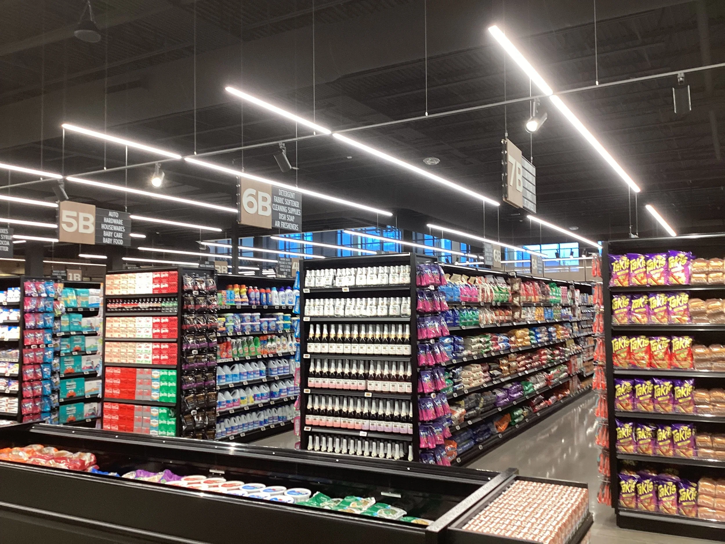 Indoor view of a grocery store aisle with black shelves stocked with snacks, drinks, and packaged goods, under modern linear ceiling lights.