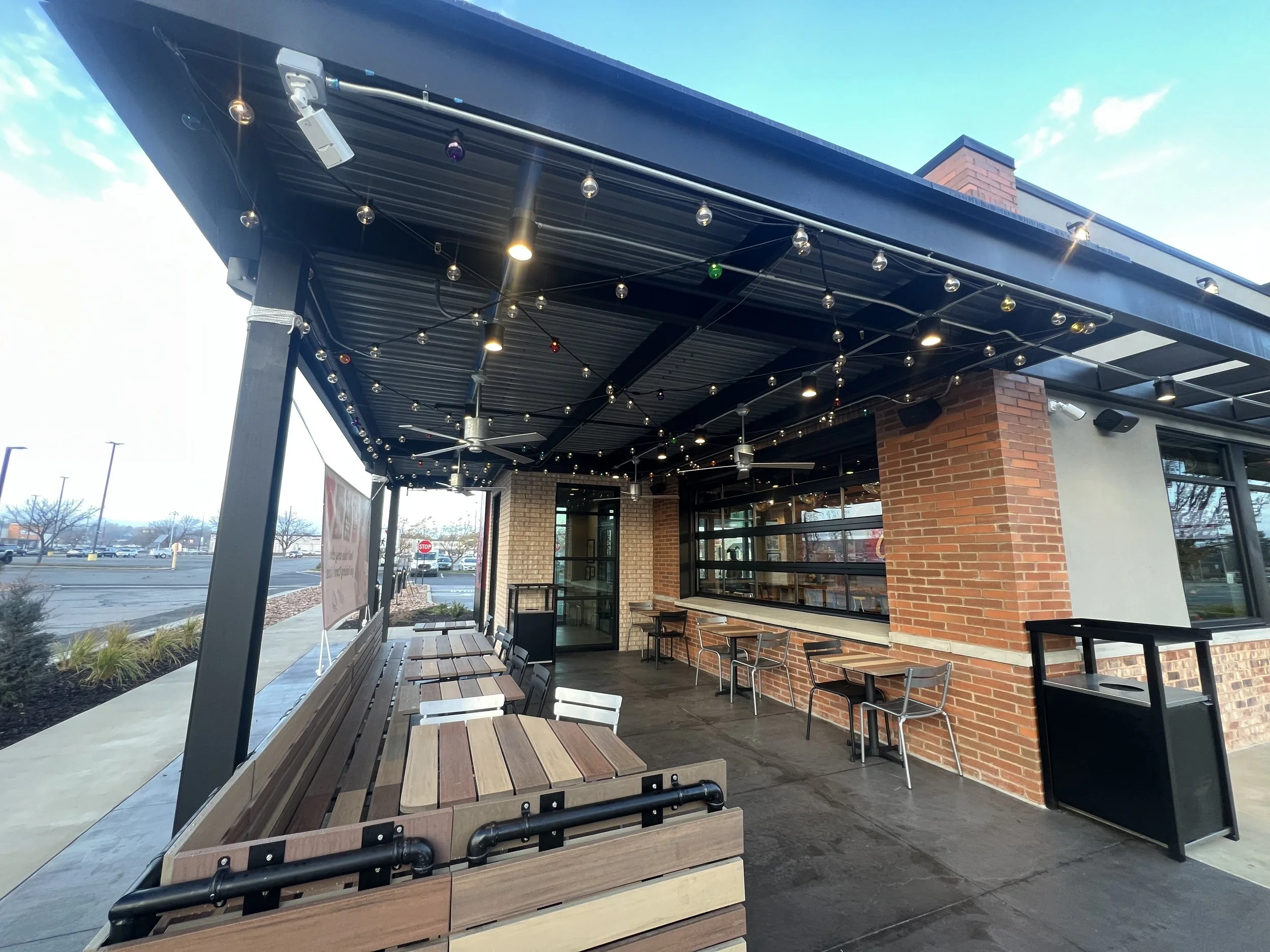 Empty outdoor patio of a restaurant with wooden tables and chairs, string lights, ceiling fans, and a brick wall, overlooking a parking lot.