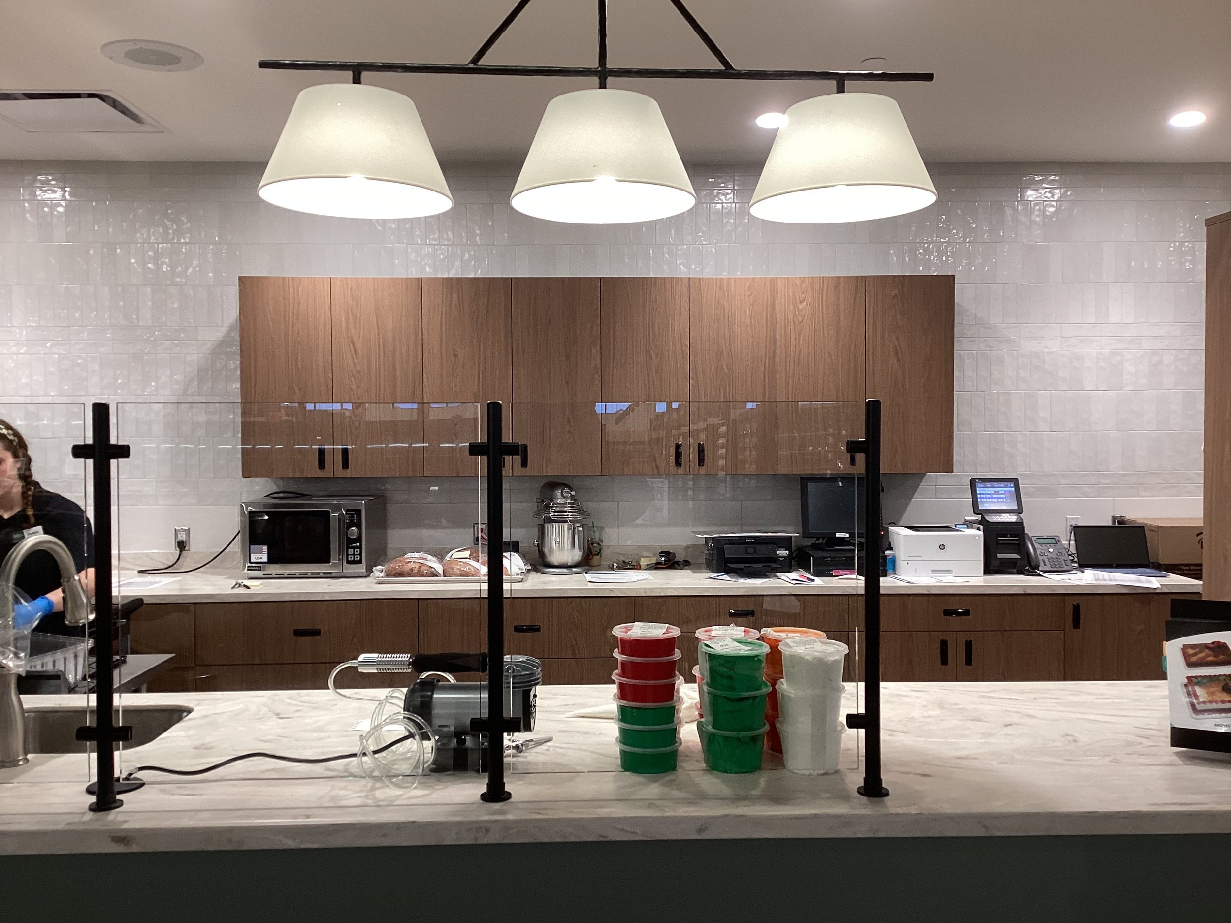 A modern bakery or cafe counter with mesas in the background, with stacks of red, green, and white containers on the counter in the foreground, and various kitchen appliances, including a microwave, stand mixer, printer, and cash register, on the back counter.