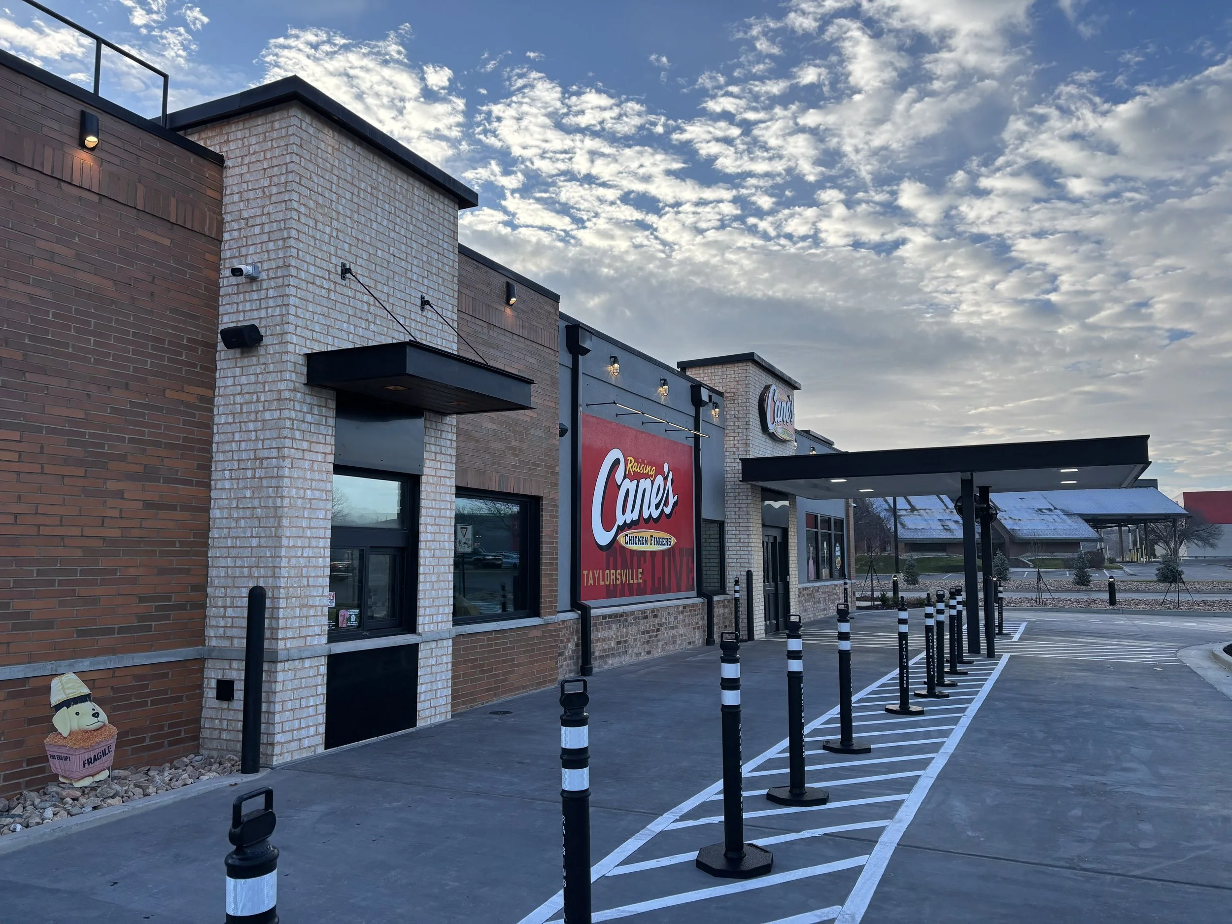 Empty fast food restaurant parking lot with black and white striped poles and a covered drive-thru lane under a partly cloudy sky.