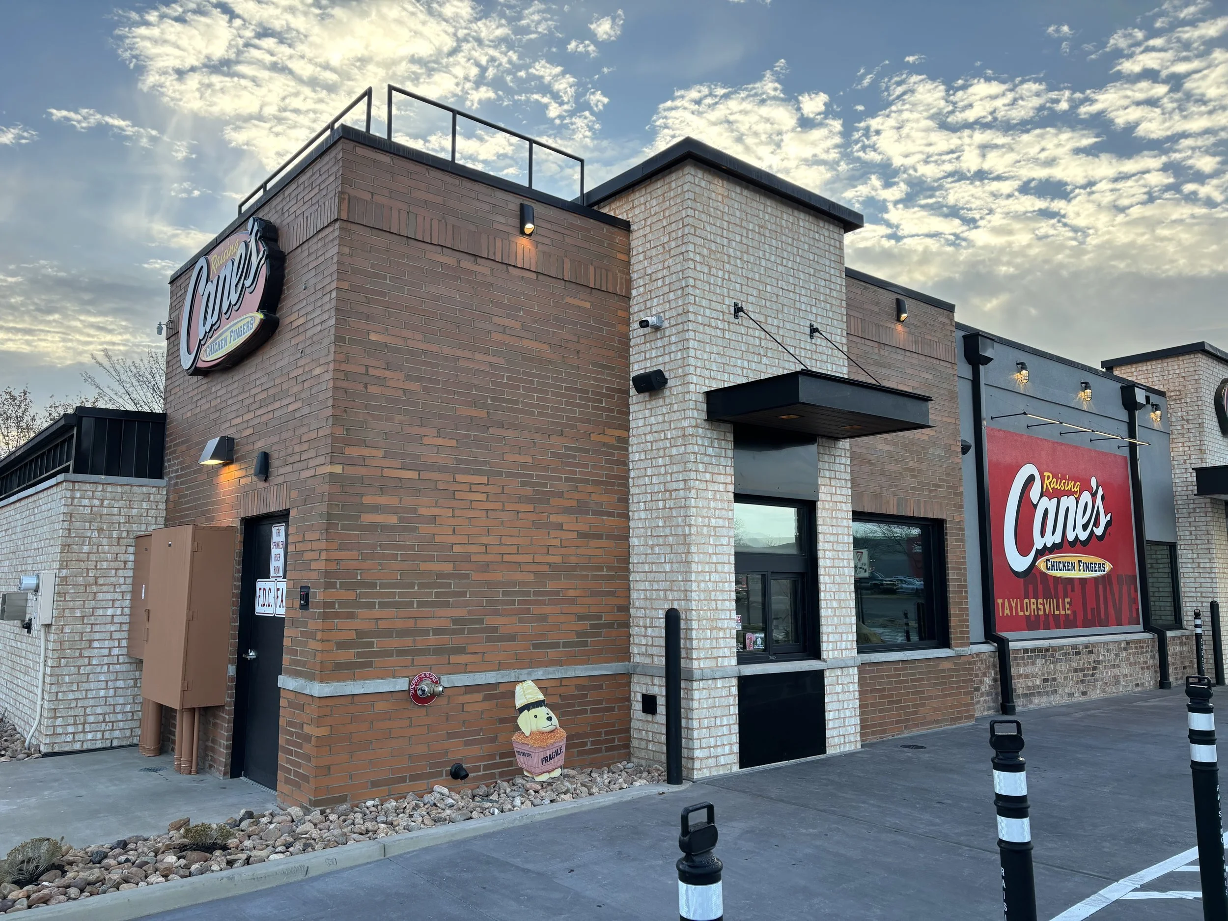 Exterior view of Raising Cane's restaurant with brick facade, neon sign, large sign on the wall, and parking lot, during cloudy daytime.