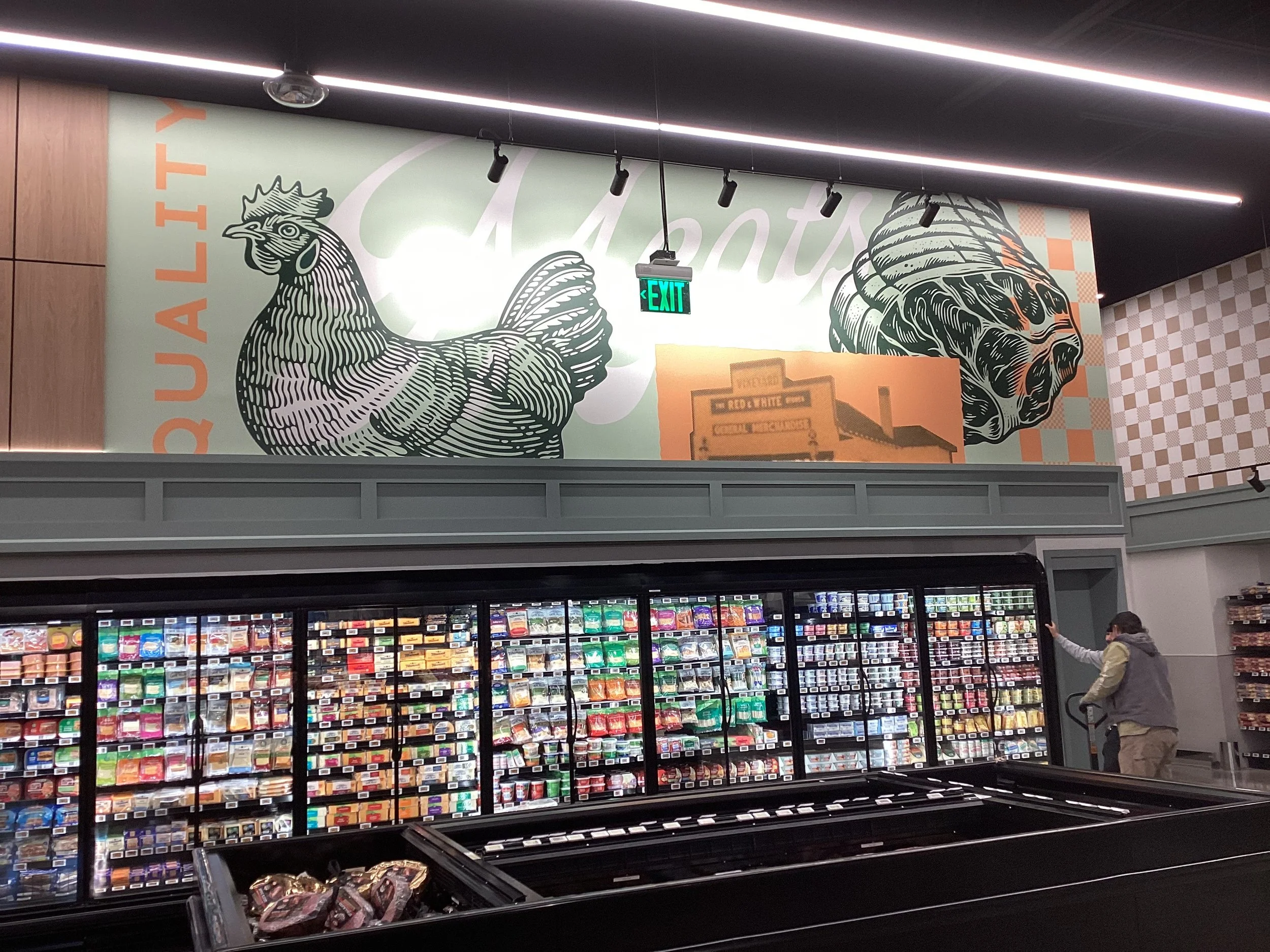 Inside a grocery store with a large mural of a chicken on the wall, printed in black and white, and an exit sign hanging from the ceiling. A person wearing a jacket and mask is reaching for an item on the refrigerated shelves filled with various chee