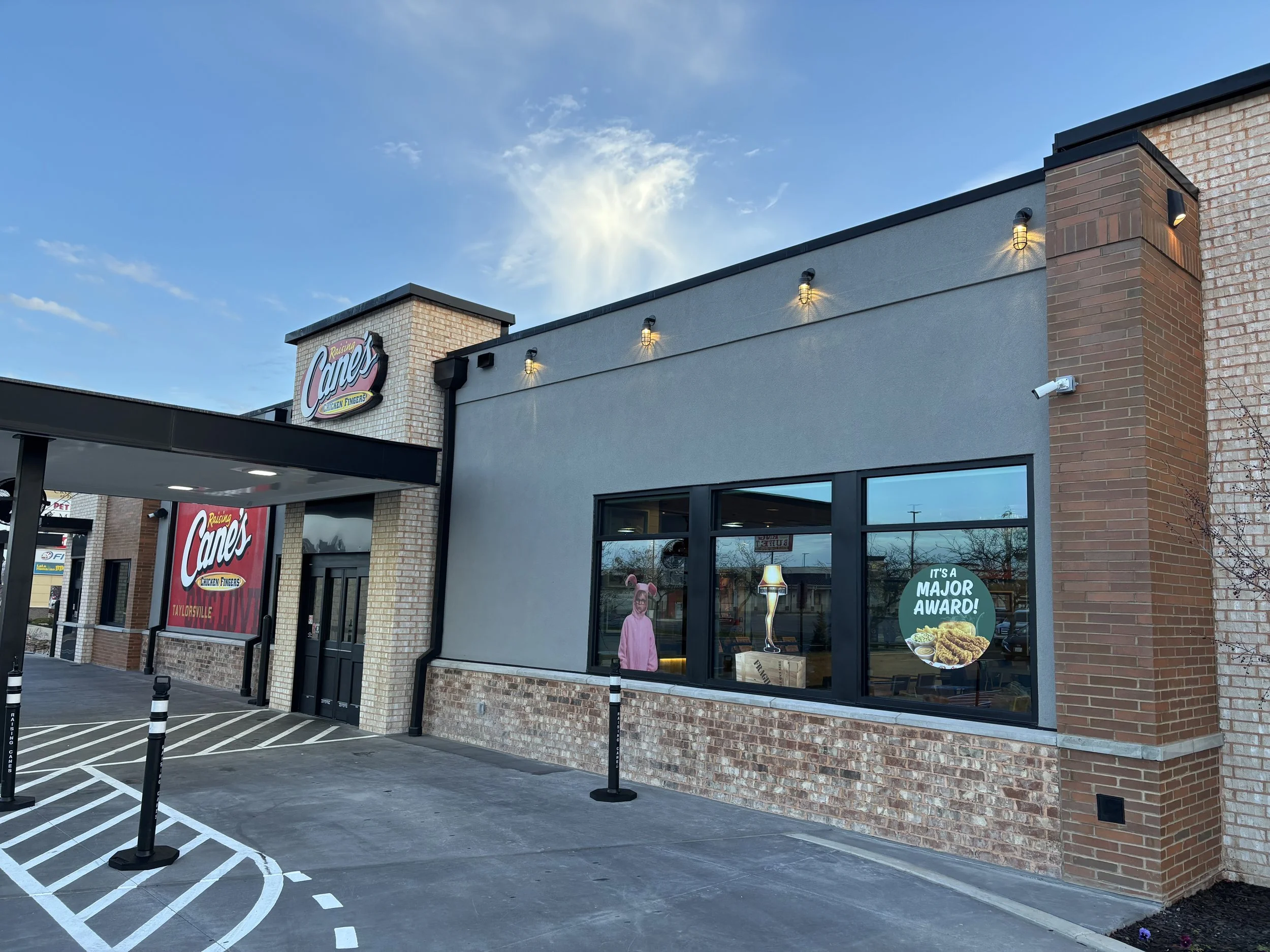 Exterior view of Raising Cane's Chicken Fingers restaurant with signage, large window, and a parking lot with striped parking spaces. The sky is clear with some clouds.