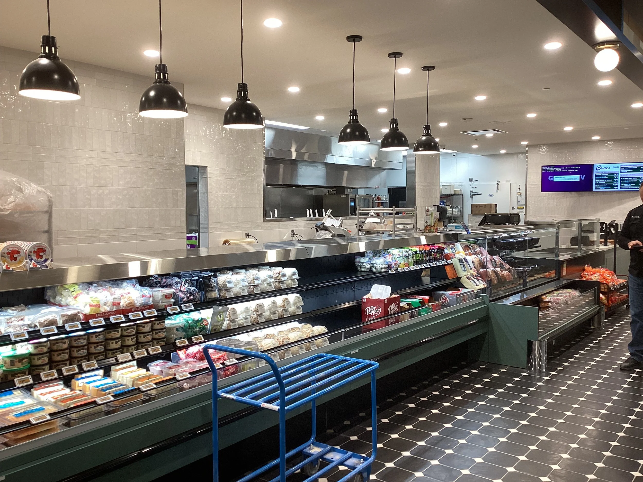 Grocery store cheese section with packaged cheeses, snacks, and a blue shopping cart in front.