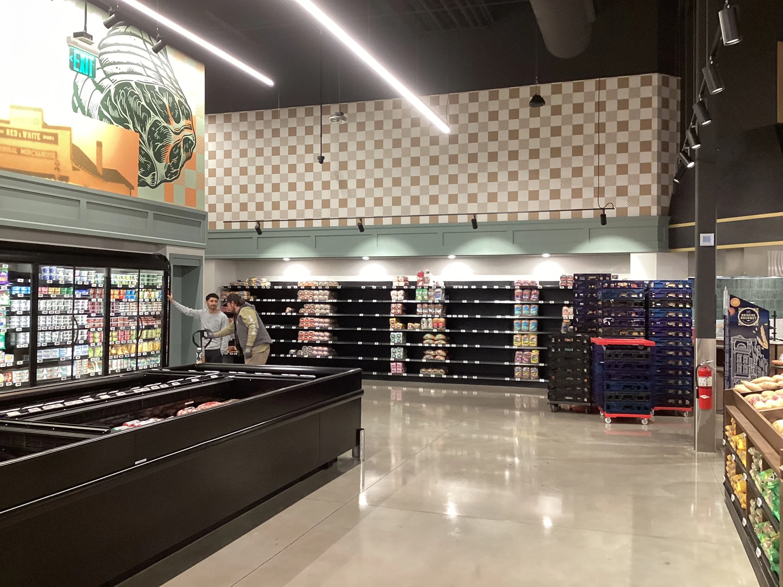 Inside a grocery store with empty shelves, a man shopping and a store employee near refrigerated section, with black freezer units, stacked crates, and a colorful mural on the wall