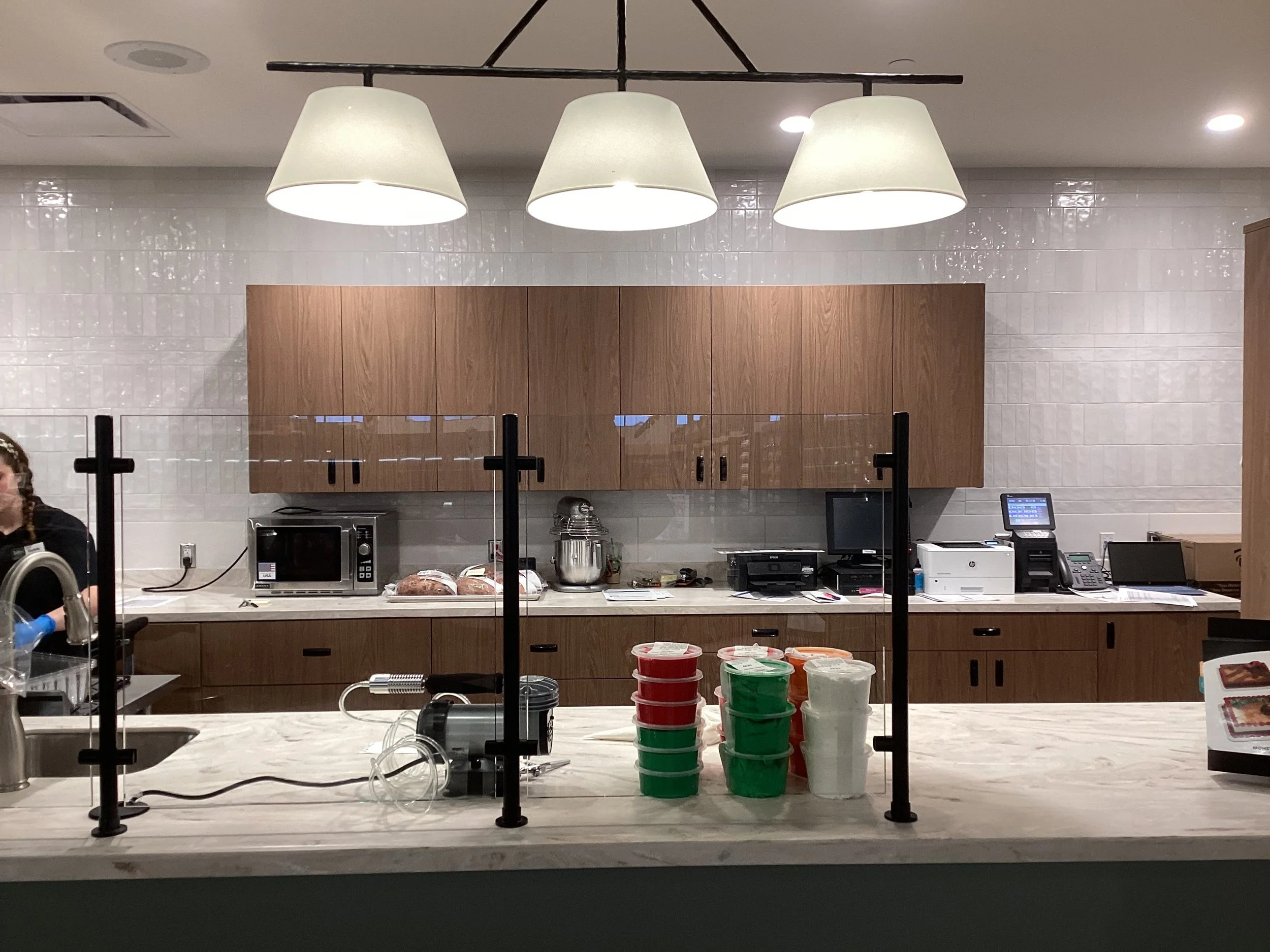 View of a commercial kitchen countertop, various kitchen appliances, and a person on the left. The background has wooden cabinets and a white tiled wall, with three large ceiling pendant lights hanging above.