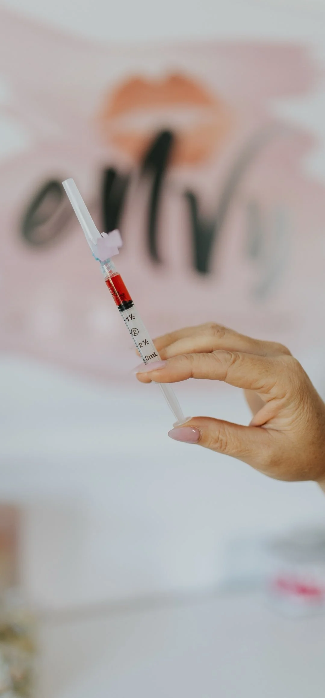 Close-up of a person's hand holding a syringe with a red liquid, with a blurred tattoo of lips and words in the background.
