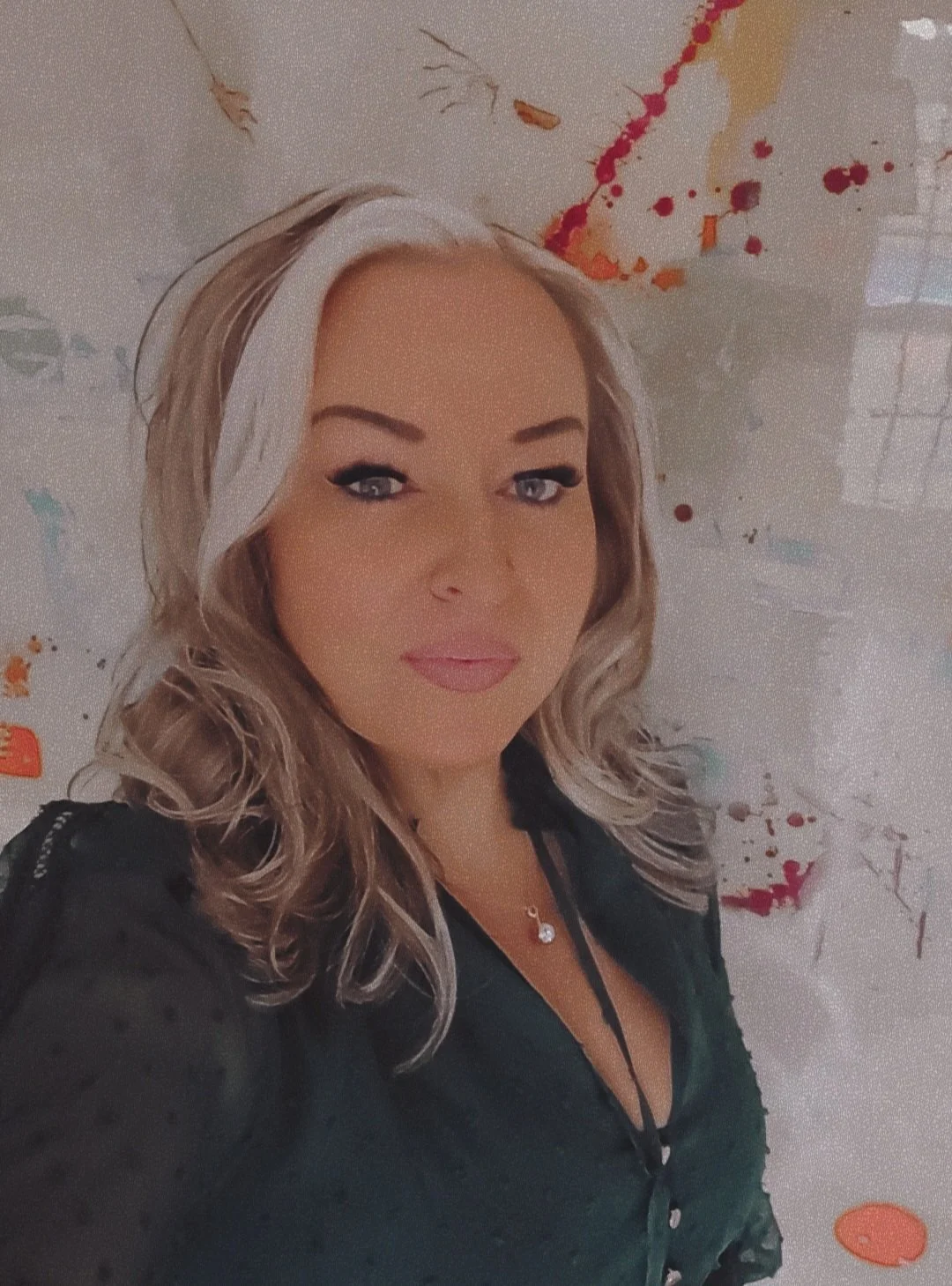 A woman with wavy blonde hair and light makeup taking a selfie in front of a colorful abstract background with red, orange, and gold paint splatters.