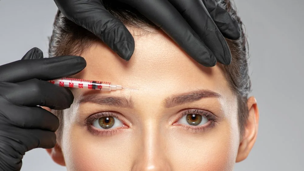 A woman with light skin and hazel eyes receiving a cosmetic injection in her forehead from a professional wearing black gloves.