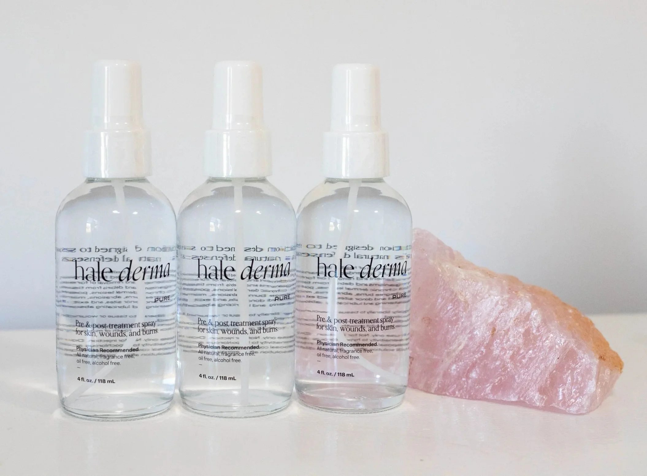 Three bottles of Hale Derma pre and post treatment spray with a pink Himalayan salt rock next to them on a white surface against a plain white background.