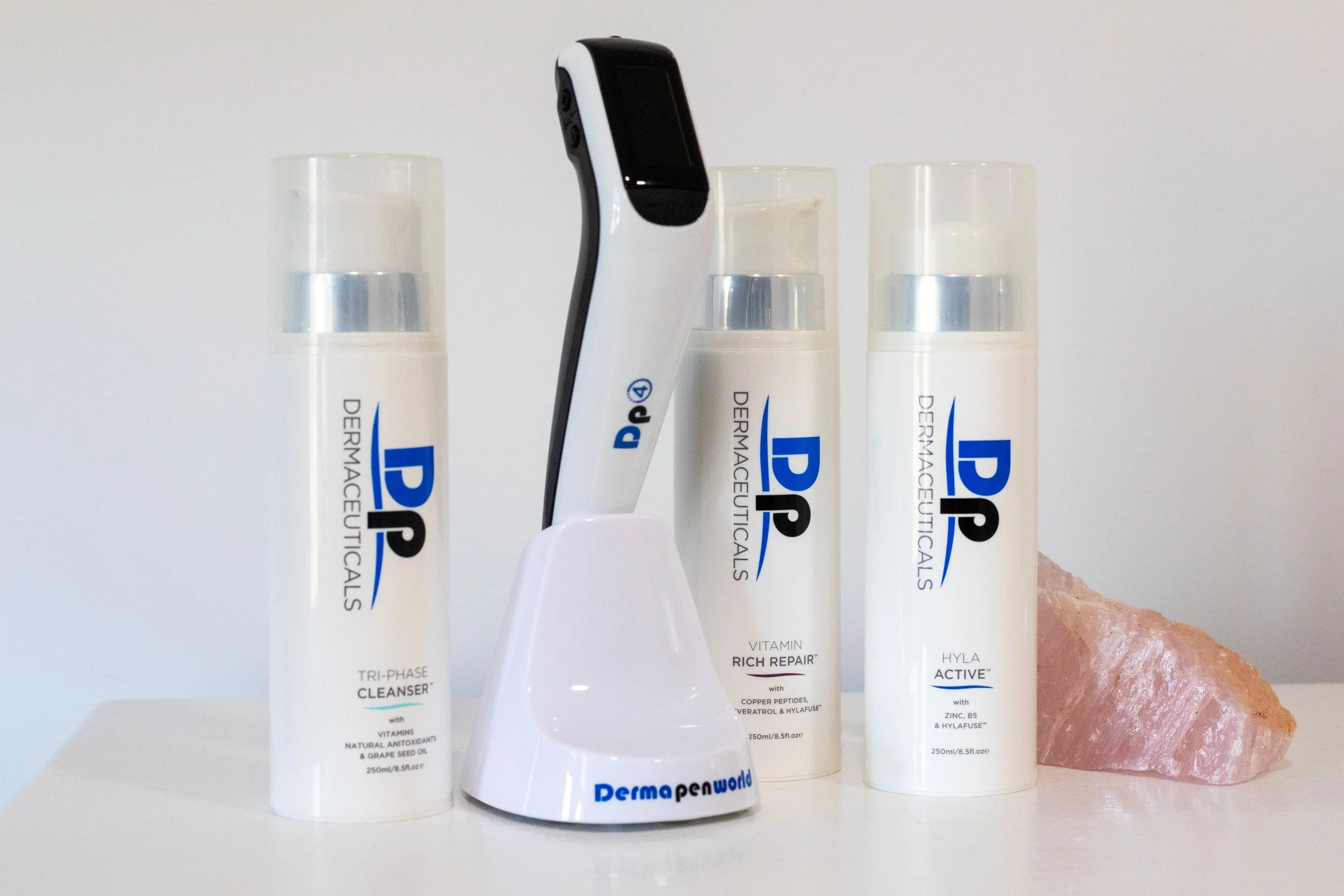 Three skincare bottles labeled Dermaceuticals and a black and white electronic device in a white stand, all on a white surface with a pink crystal-like object beside them.