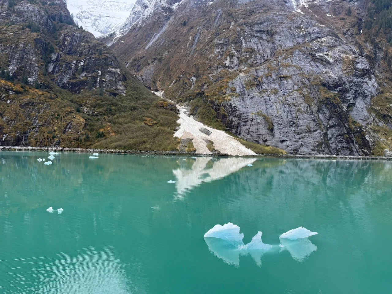 A glacier-fed lake with floating ice chunks set against steep, rocky mountains with some snow and sparse vegetation.