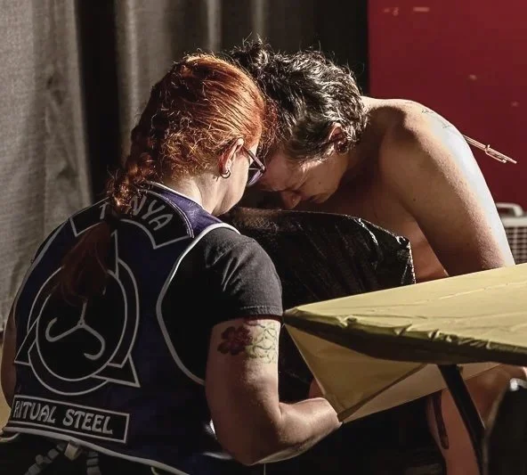 A woman with red hair and glasses is comforting a man who has just been pierced. He is shirtless and they are  in a dimly lit room.
