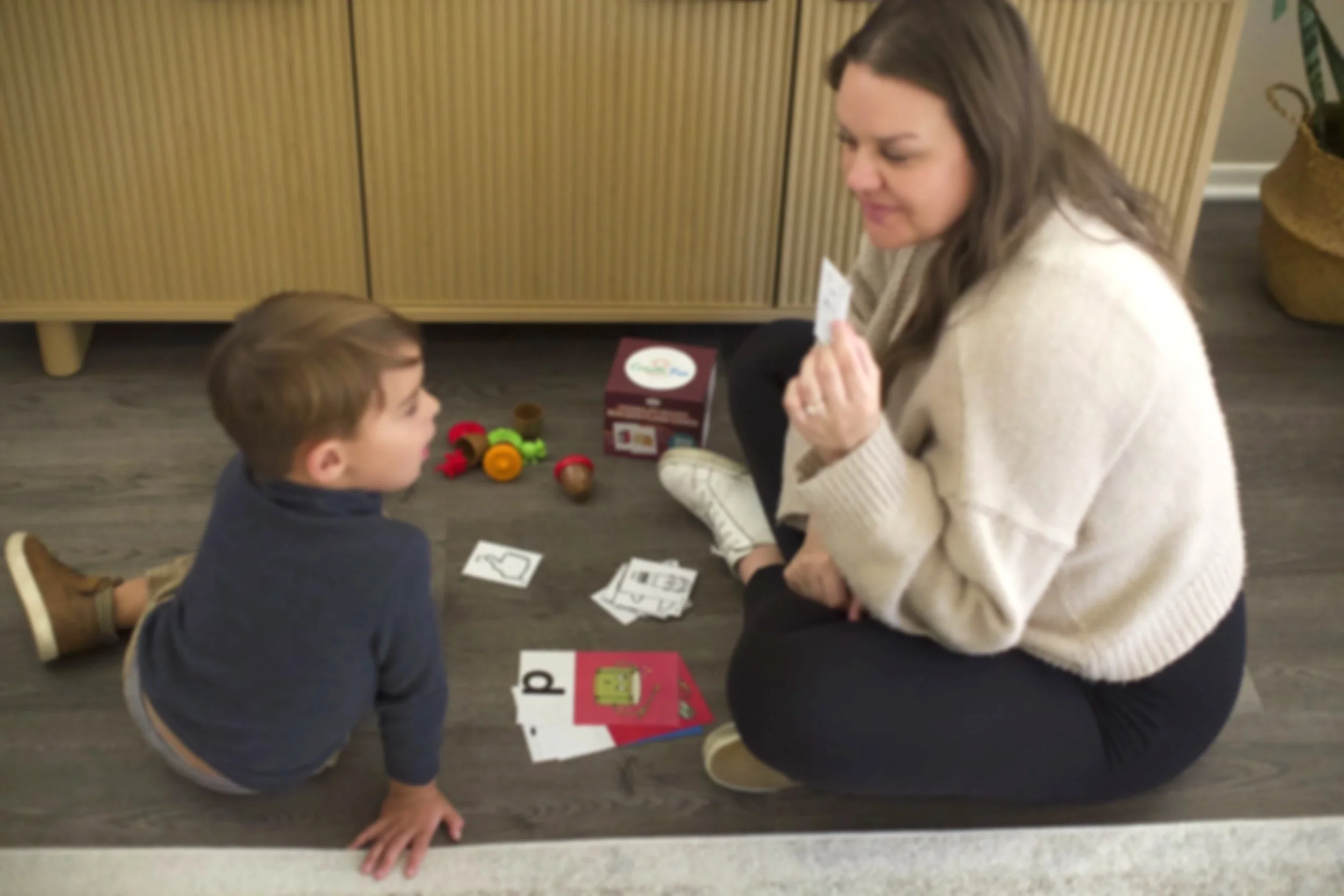 A woman kneeling on the floor playing a card game with a young boy. There are various game pieces and cards spread on the floor between them, with a box of game pieces behind them. The woman is wearing a cream-colored sweater, and the boy is in a dark hoodie with brown hair.