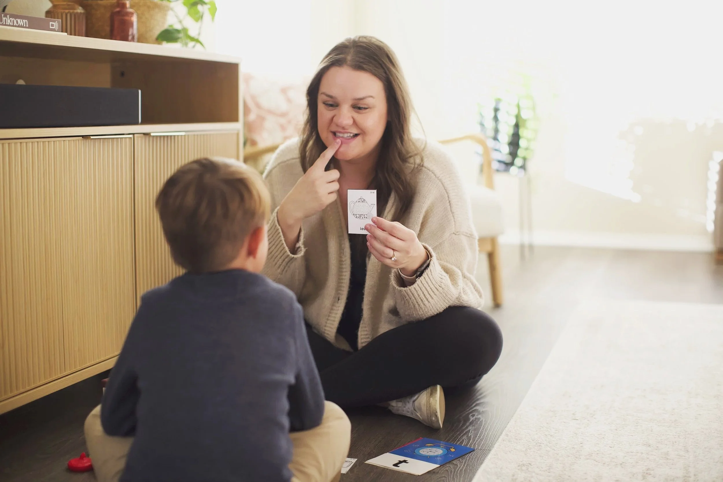 A teacher sitting on the floor, showing a picture card to a young boy during an educational activity. The setting is bright and cozy, with shelves and a potted plant in the background.