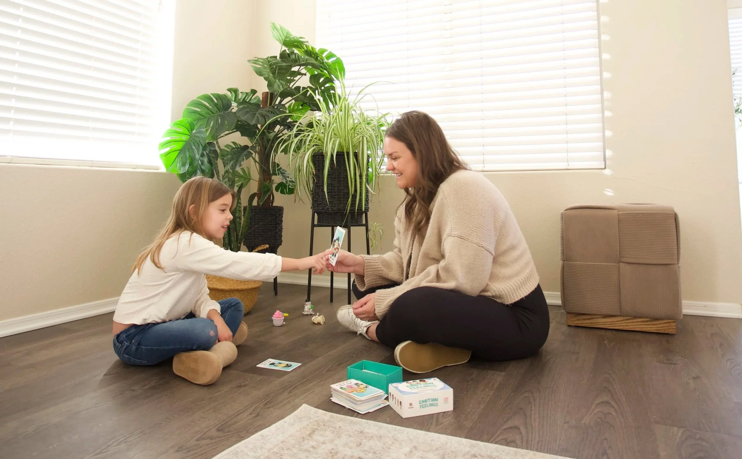 A woman and a girl sitting on the wooden floor playing a card game together in a bright room with large windows, indoor plants, and a cardboard box of cards and game pieces.