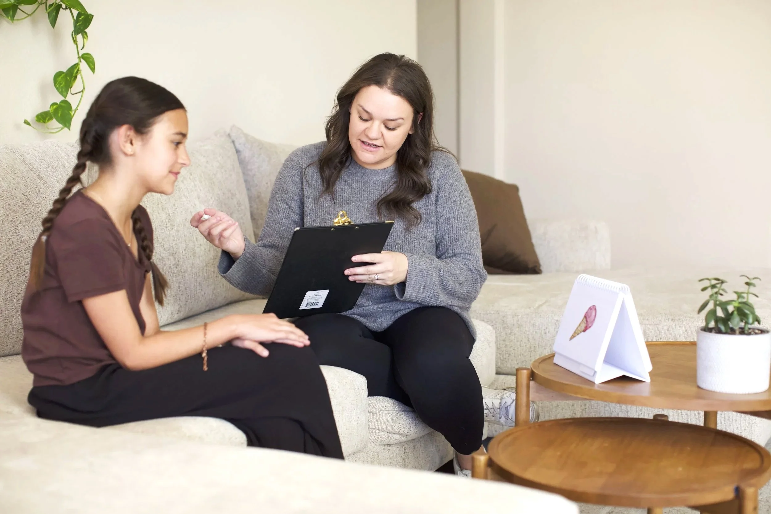 Adult woman with dark brown hair talking to young girl with braided brown hair while looking at a clipboard, sitting on a beige couch in a room with light-colored walls and a small wooden coffee table with a framed ice cream painting, potted plant, and a standee in the background.