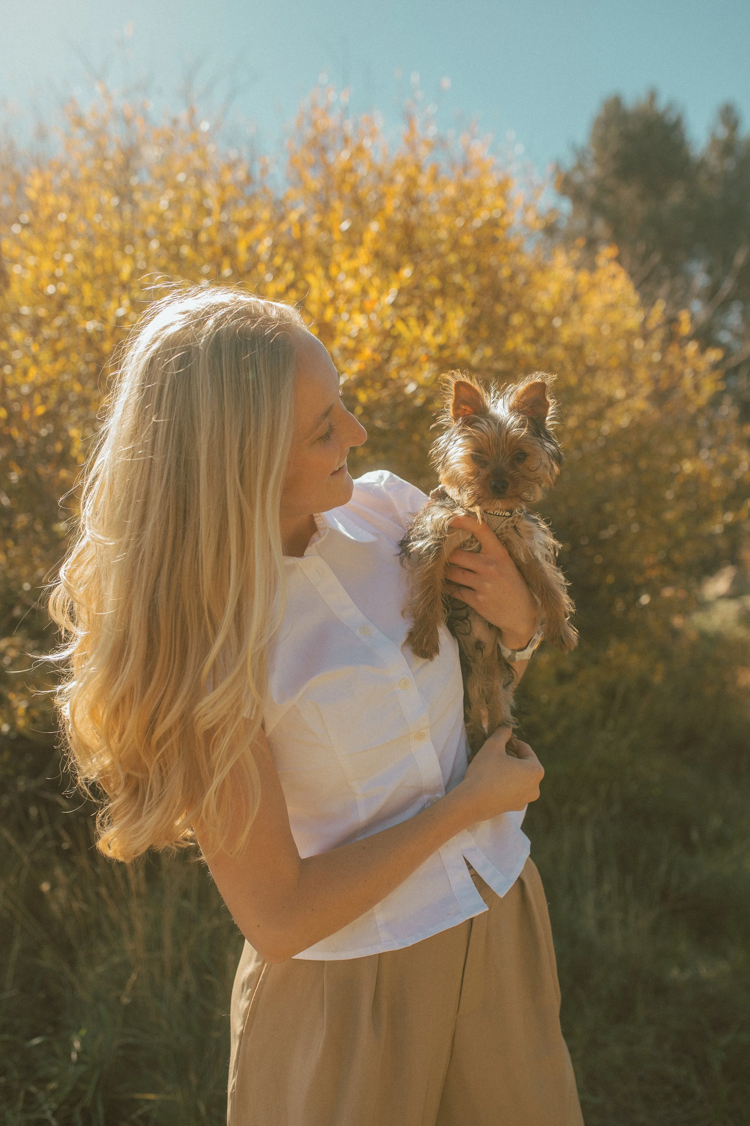 A woman with long blonde hair holding a small dog outdoors during fall, with trees with golden leaves in the background.