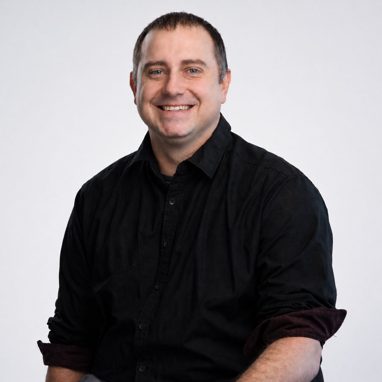 Meet our Quality Assurance Administrator, Kenny Robertson. A man with a beard and short hair smiling outdoors on a grassy hill with a cloudy sky and green hills in the background, wearing a lavender polo shirt and khaki pants.