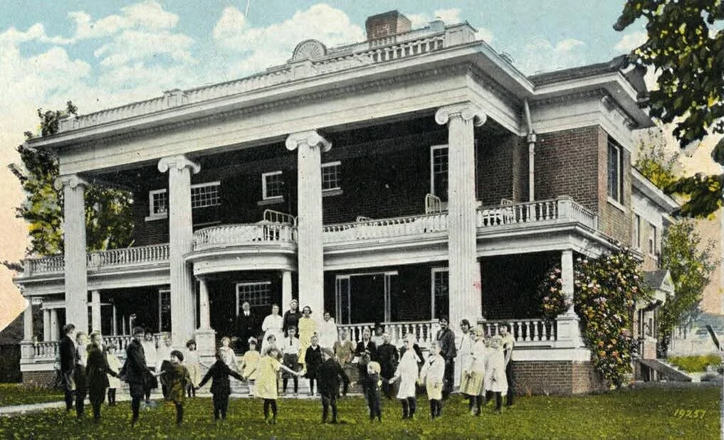 Group of children and adults standing in front of a historic, two-story mansion with white columns and a wraparound porch.