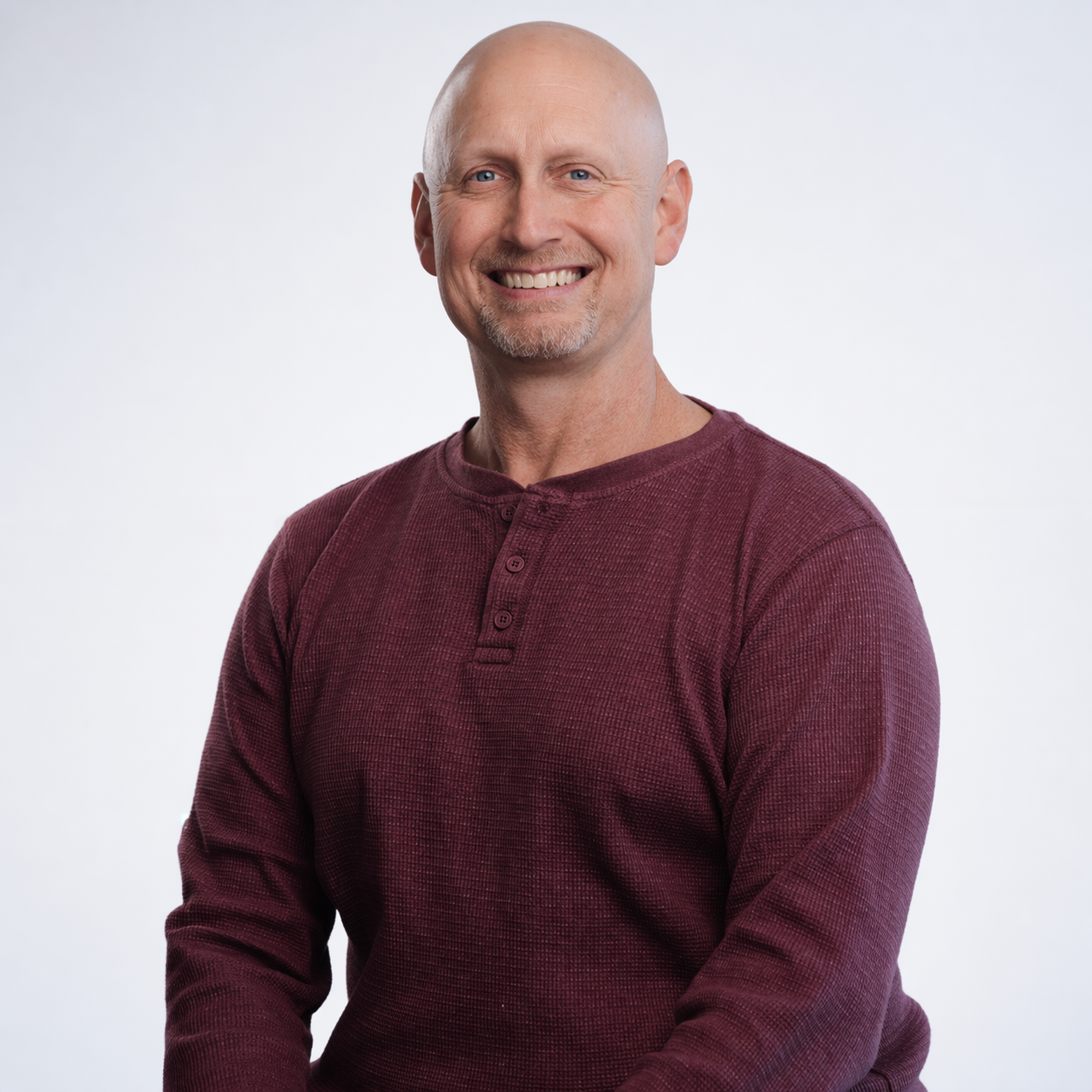 Meet our executive director, Brian Pope. A smiling man with a bald head and goatee, wearing a navy blue short-sleeve button-up shirt and matching navy pants, standing outdoors with green hills and cloudy sky in the background.
